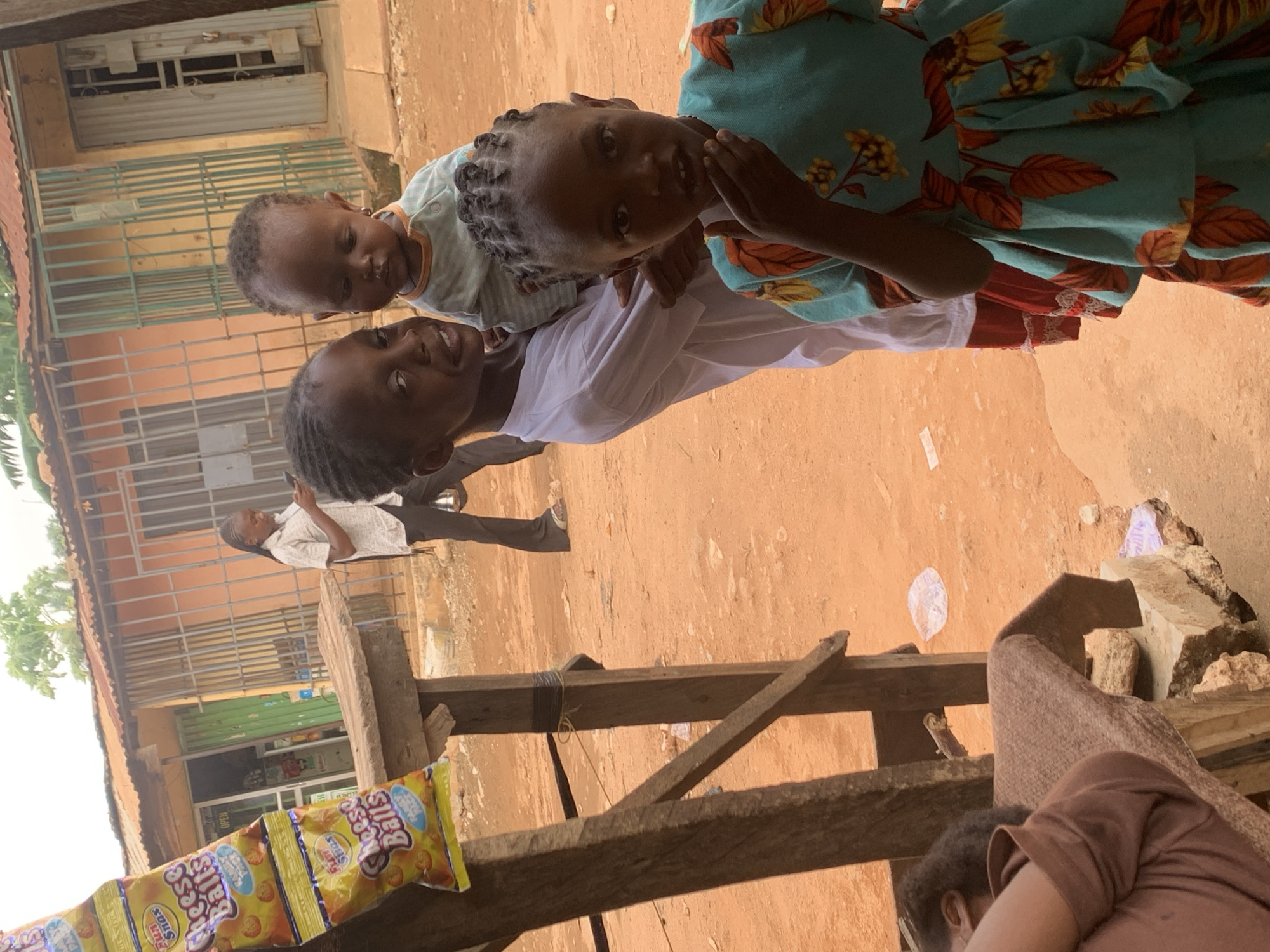 The image shows a scene in Sabon Tasha, Nigeria. Three children are the focus, with one older child carrying a younger one on their back. A third child, wearing a dress with a floral pattern, is to the right. They appear to be looking directly at the camera. In the background, a man in a light-colored shirt is standing in front of a shop. A wooden structure is in the foreground, with packages of "Cheese Balls" snacks visible on it. The ground is dirt and the buildings appear to be simple, possibly made of concrete or cinder blocks.