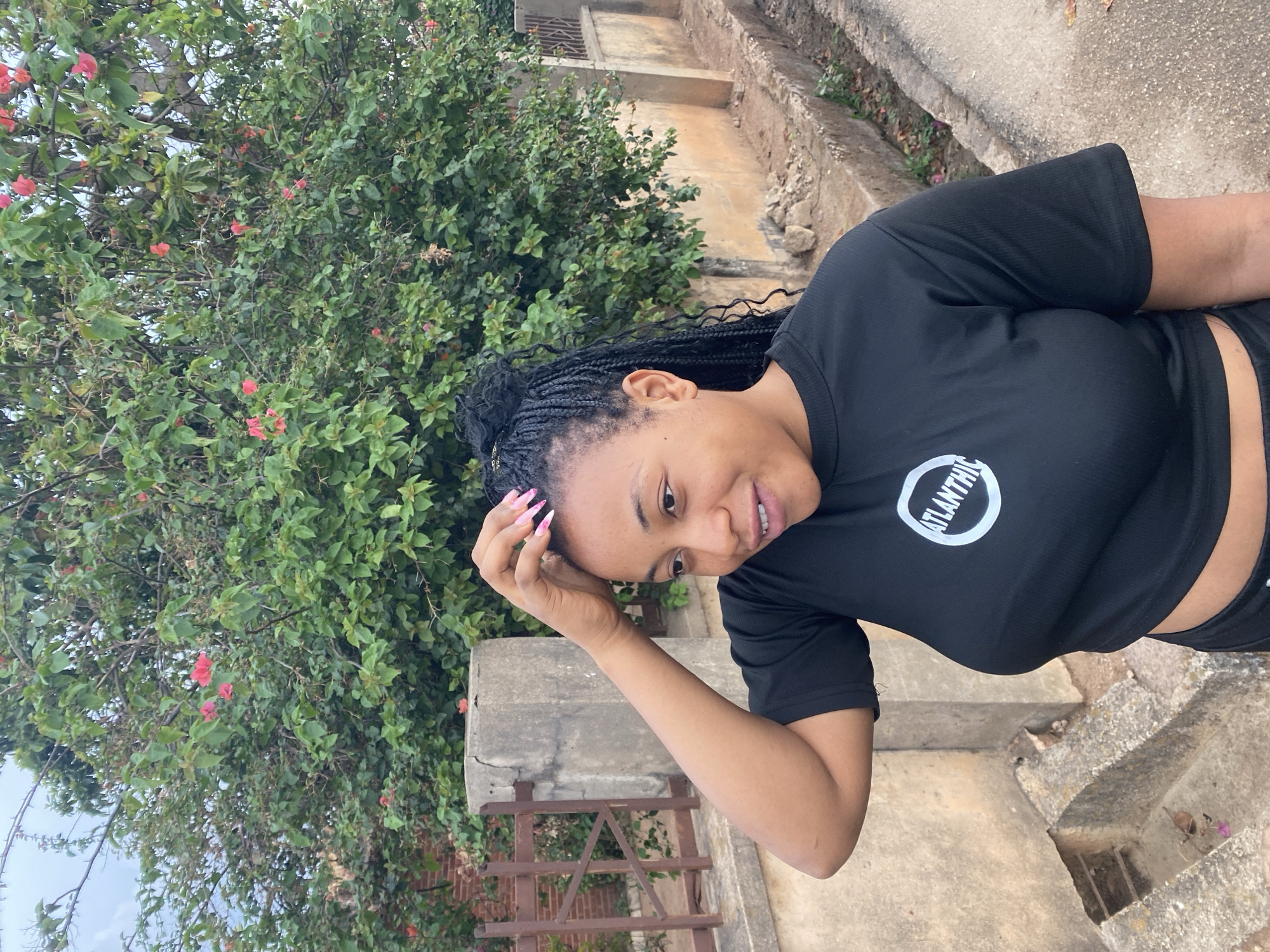 A woman with braided hair is standing outside near some steps and a concrete wall. She is wearing a black shirt and shorts. A tree with green leaves and red flowers is behind her. The location is Jos, Nigeria.