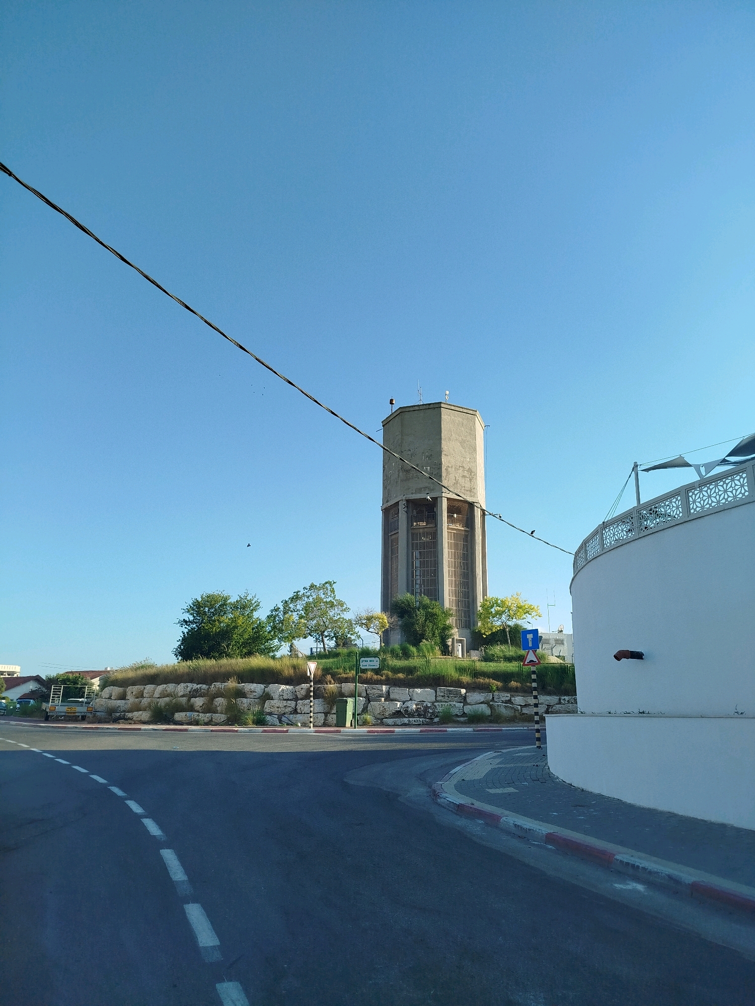 A tall, hexagonal concrete water tower stands prominently in the background of this outdoor scene, its weathered surface suggesting age. The tower is situated on a grassy hill, surrounded by trees and shrubs, and supported by a stone wall. In the foreground, a curved asphalt road with white dashed lane markings leads the viewer's eye towards the tower. To the right of the road, a white circular building with an ornate railing is partially visible, casting a shadow on the street. The sky above is a clear, bright blue, with a black wire stretching diagonally across the upper portion of the frame. A few birds can be seen flying in the distance. The scene appears to be a street in the city of Ashkelon, Israel, with elements of both urban infrastructure and natural landscaping.