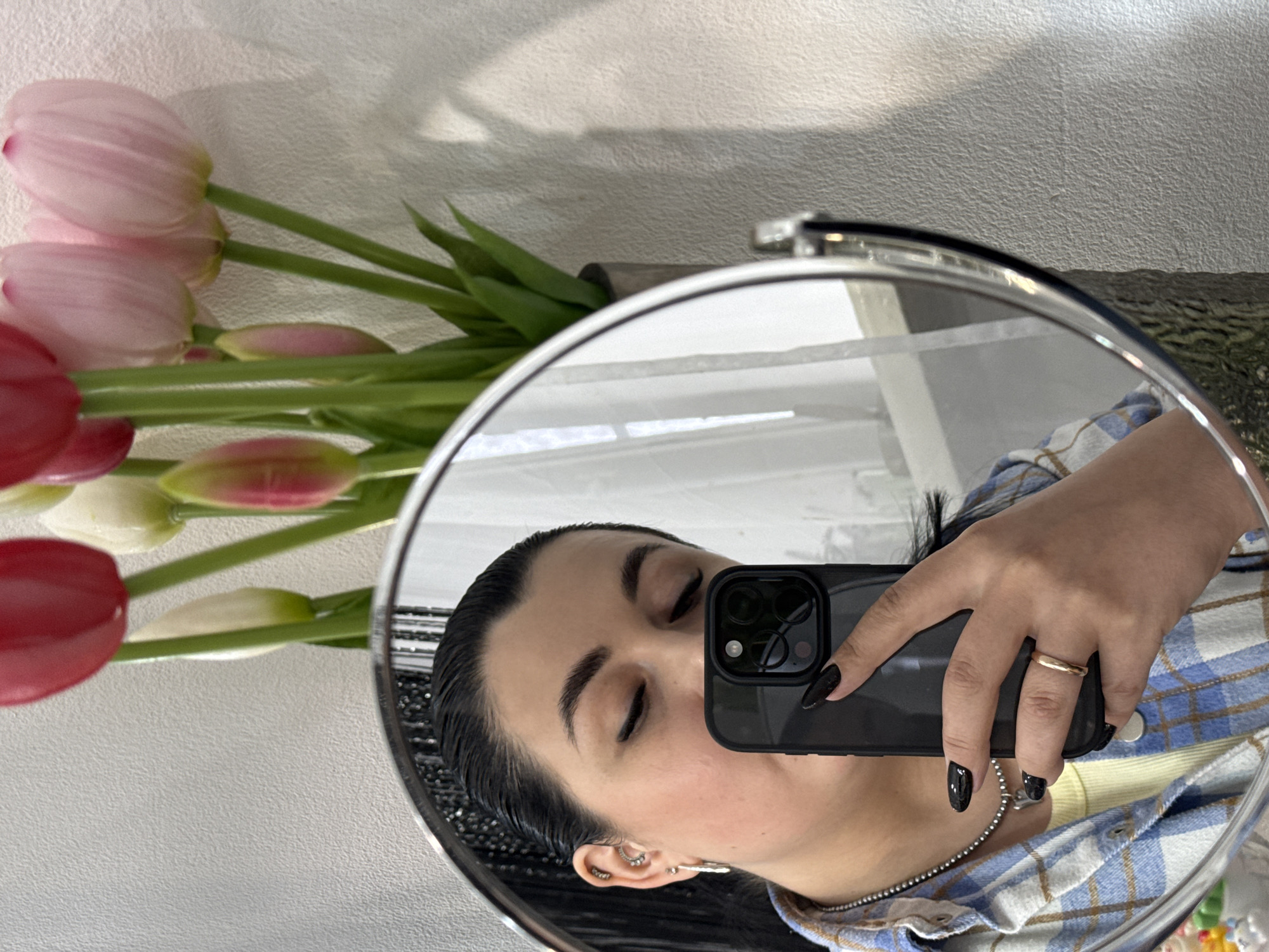 A woman is reflected in a circular mirror while taking a selfie with a black smartphone. She has dark hair, pale skin, and is wearing a blue and white plaid shirt with a yellow undershirt. Her nails are painted black, and she wears a gold ring on her right hand. A bouquet of tulips in shades of pink and red, along with green leaves, sits to the left of the mirror. The background appears to be a wall with a light texture. The location is unknown and cannot be determined from the image.