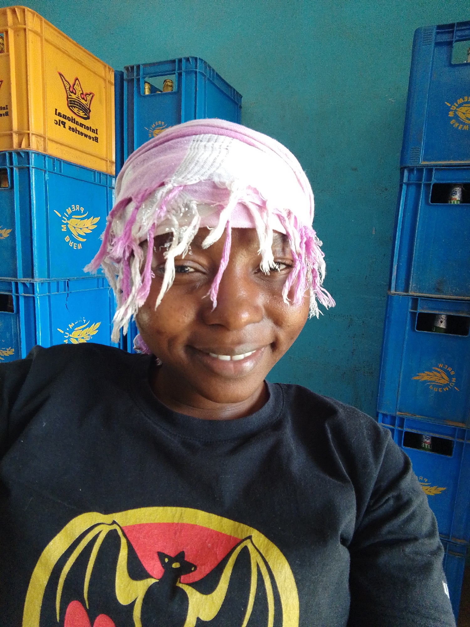 A smiling woman wears a pink and white headscarf and a black t-shirt with a yellow and red Bacardi bat logo. Stacks of blue and yellow beer crates are visible in the background, next to a teal wall. Bottles can be seen inside the beer crates. The location is likely Jos, Nigeria.