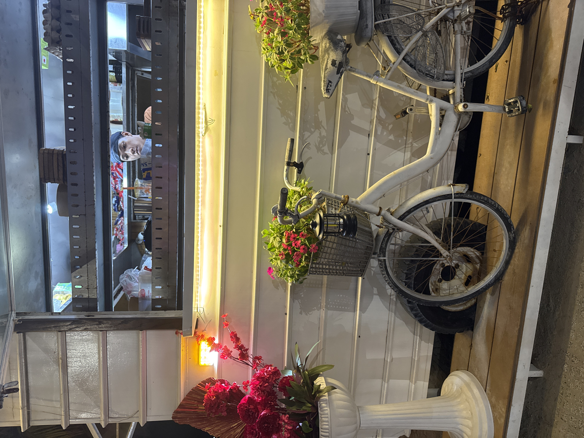 The image displays a vibrant night scene, rotated 90 degrees counter-clockwise, showing the facade of a shop or stall on the right and a glimpse of its interior on the left.
On the right side, a white, vintage-style bicycle is mounted vertically against a white wall constructed from horizontal planks. The bicycle shows signs of age and wear, with some rust visible on its metallic parts like the spokes and pedals. Baskets are attached to the handlebars and potentially over the rear wheel. Both the handlebar basket and an area near the bicycle's seat hold pots of green plants with small, bright pinkish-red flowers, serving as decoration.
Running vertically along the edge of the white planked wall, beside the shop's opening, are two bright yellow LED strip lights, illuminating the facade. Below the lower strip light, a small, amber-colored rectangular light fixture is mounted. Beneath this light, a large, ornate white planter with a fluted design holds a prominent arrangement of artificial red flowers and fan-shaped, golden-brown decorative elements. The ground beneath this area transitions from wooden planks to a darker, possibly concrete or paved surface.
On the left side of the image, through a rectangular opening or window, the interior of the shop is visible. A man with a dark blue or black cap and a light-colored, possibly plaid shirt, is looking out towards the viewer. His expression is neutral. Behind him, the shop's interior is brightly lit, revealing shelves stocked with various products, including packaged snacks and drinks. A plastic cup with a straw is visible on a counter. The opening is framed by dark gray, perforated metal shelving units or supports. To the immediate left of the opening, some stacked brown items, possibly cardboard boxes or wooden blocks, are present.
The artificial lighting strongly suggests the photograph was taken in the evening or at night, creating an inviting and decorative atmosphere for the establishment. No significant text is clearly visible.