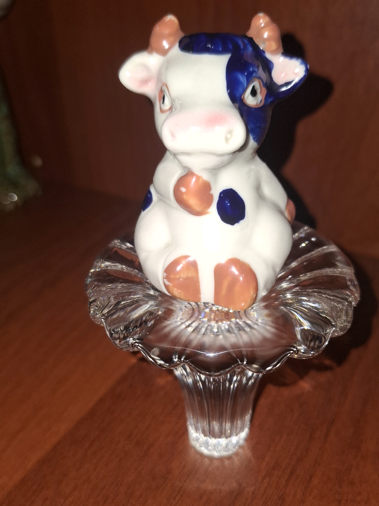 A close-up, indoor, eye-level shot features a small, white ceramic cow figurine with blue and orange markings, sitting on a clear, fluted glass pedestal. The cow has large, pink ears and a small pink snout. Its blue markings are on its head and back, while its orange markings are on its hooves and two spots on its body. The glass pedestal has a petal-like top and a ribbed stem. The background is a dark, reddish-brown wooden surface, likely a table or shelf. A shadow of the figurine is cast on the surface behind it. The scene is lit by artificial light, creating highlights on the ceramic and glass. There are no people or animals present in the image. The location in Almaty, Kazakhstan cannot be determined from the visual information provided.