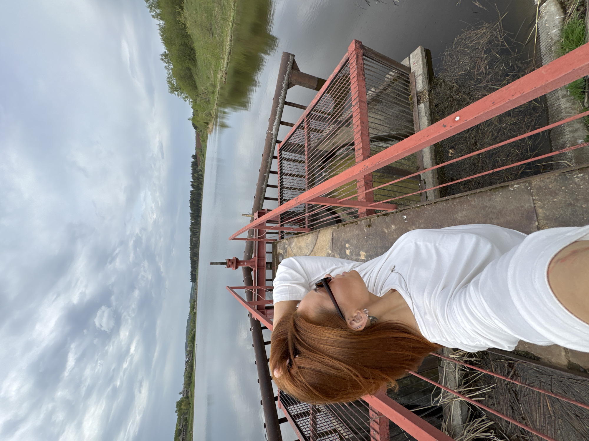 A selfie photograph shows a person with red hair, wearing a white shirt and sunglasses, standing on a metal structure, possibly a bridge or dam, with a red railing. The person is oriented such that the landscape appears vertically behind them. The background features a body of water, likely a river or lake, with trees and greenery visible on the opposite bank. The sky is overcast, suggesting a cloudy day. The location is purportedly in Nozhovka, Russia, but this cannot be verified from the image alone. The metal structure appears to be aged and weathered, with signs of rust. It provides a platform over the water, with metal grating and infrastructure visible below.