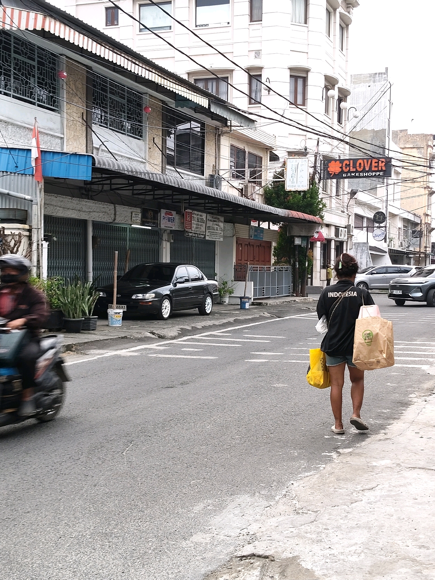 In the City of Medan, Indonesia, a street scene unfolds under an overcast sky. The foreground features a gray asphalt road with white lane markings, leading towards a sidewalk on the right. A person on a speeding motorcycle, blurred by motion, is captured on the left side of the road, wearing a dark jacket and a helmet. On the sidewalk, a woman walks away from the camera, carrying two shopping bags: a yellow one in her left hand and a larger brown paper bag in her right. She is wearing a black jacket with "INDONESIA" printed in white on the back, and denim shorts. Her footwear appears to be sandals. Behind her, a silver car is parked on the street.
The middle ground reveals a row of buildings with shops and awnings. A dark-colored sedan is parked in front of one of the establishments, partially obscured by potted plants. A white bucket with some sort of label is situated near the car. A red and white Indonesian flag is visible on the left, near a blue awning. Further down the street, a prominent sign with an orange clover leaf and the text "CLOVER BAKESHOPPE" is illuminated. Power lines crisscross above the street and buildings.
The background consists of more buildings, some of which appear to be multi-story structures, with visible windows and architectural details. The overall impression is a typical urban street in Indonesia, with a mix of commercial activity and daily life.