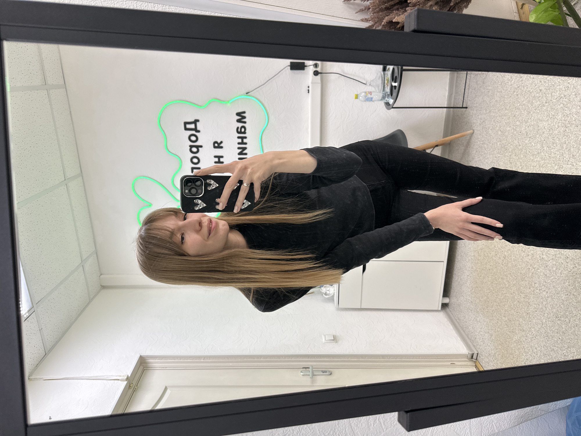 A young woman is taking a selfie in a mirror in what appears to be an office or commercial space in Chaikovsky, Russia. She is wearing a black top and black pants. The mirror is reflecting a white wall with a green neon sign that includes Cyrillic text. In the background, there is a table with items on it, a chair, a white cabinet, and a door. The ceiling has a grid pattern. The floor appears to be carpeted with a textured design.