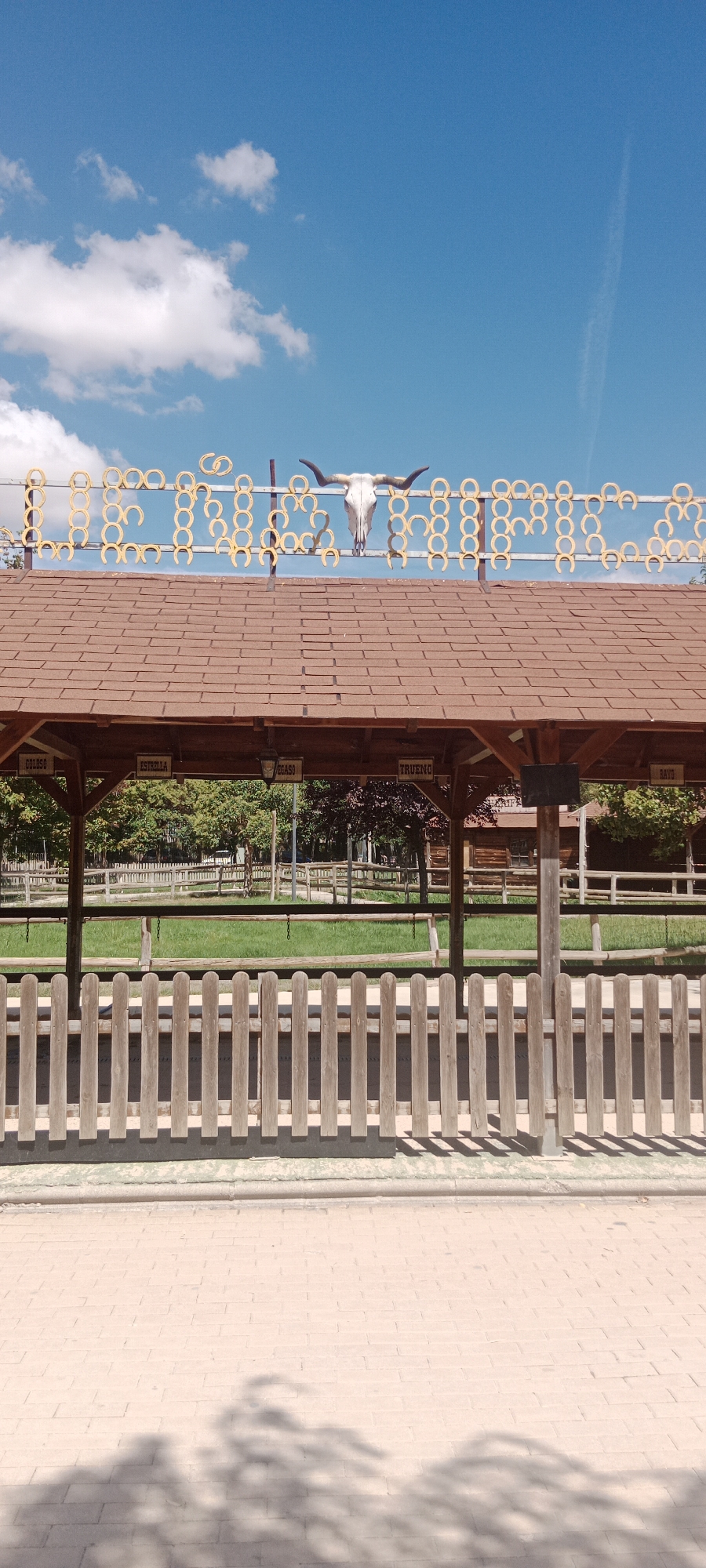 The image shows a rustic wooden structure with a brown shingled roof, likely a welcome station or an entrance to an attraction. Above the roof, a metal sign is adorned with numerous golden horseshoes and a bull skull, suggesting a western or rodeo theme. Beneath the eaves of the structure, five small wooden signs with text are hanging, with the visible signs reading "GOLOSO", "ESTRELLA", "REGASO", "TRUENO", and "RAYO". The structure is set against a backdrop of green grass and trees, with a clear blue sky and scattered white clouds above. A wooden picket fence runs along the front of the structure, separating it from a paved walkway in the foreground. The overall scene appears to be outdoors, in a park or recreational area. The location is specified as Torrejón de Ardoz, Spain. There are no people visible in the image.