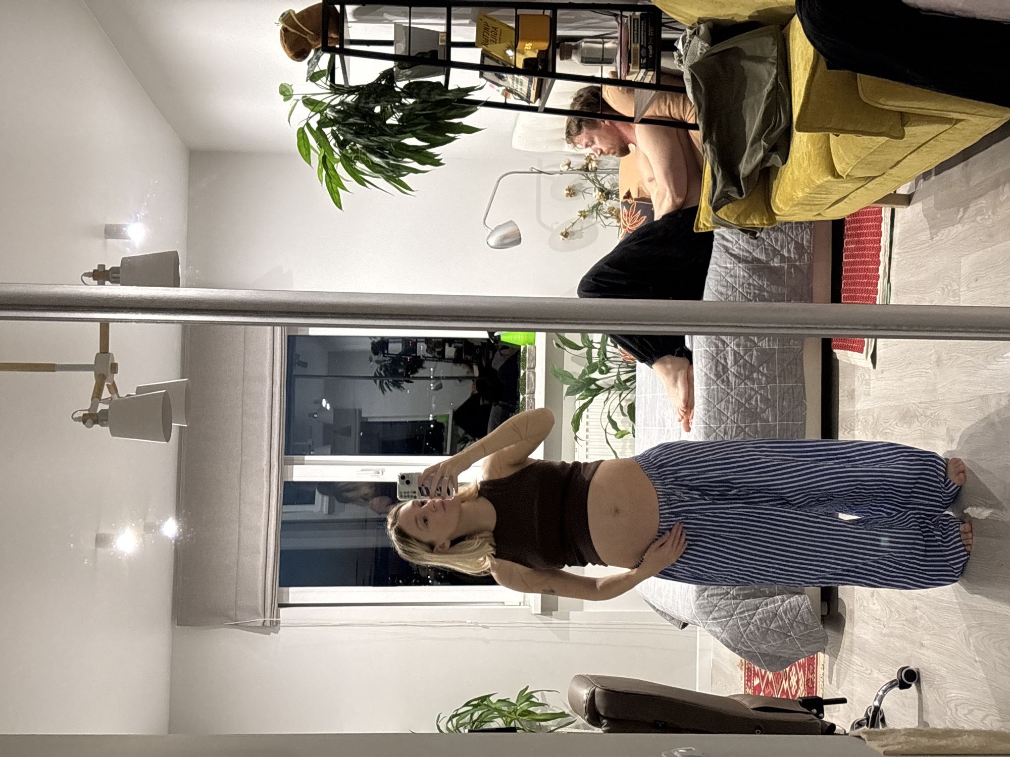 A pregnant woman stands in a bedroom, taking a mirror selfie. She is wearing a dark brown tank top and blue and white striped pajama pants. Her left hand rests on her stomach. The woman has blonde hair and is looking at her phone. In the background, a man is lying on a bed, looking towards the woman. He is shirtless and wearing black pants. A large mirror reflects the scene. The bedroom is furnished with a bed, a yellow sofa, a chair, and a bookshelf. Plants are visible in pots around the room. The overall lighting suggests it is nighttime. The location is identified as Nizhny Novgorod, Russia.