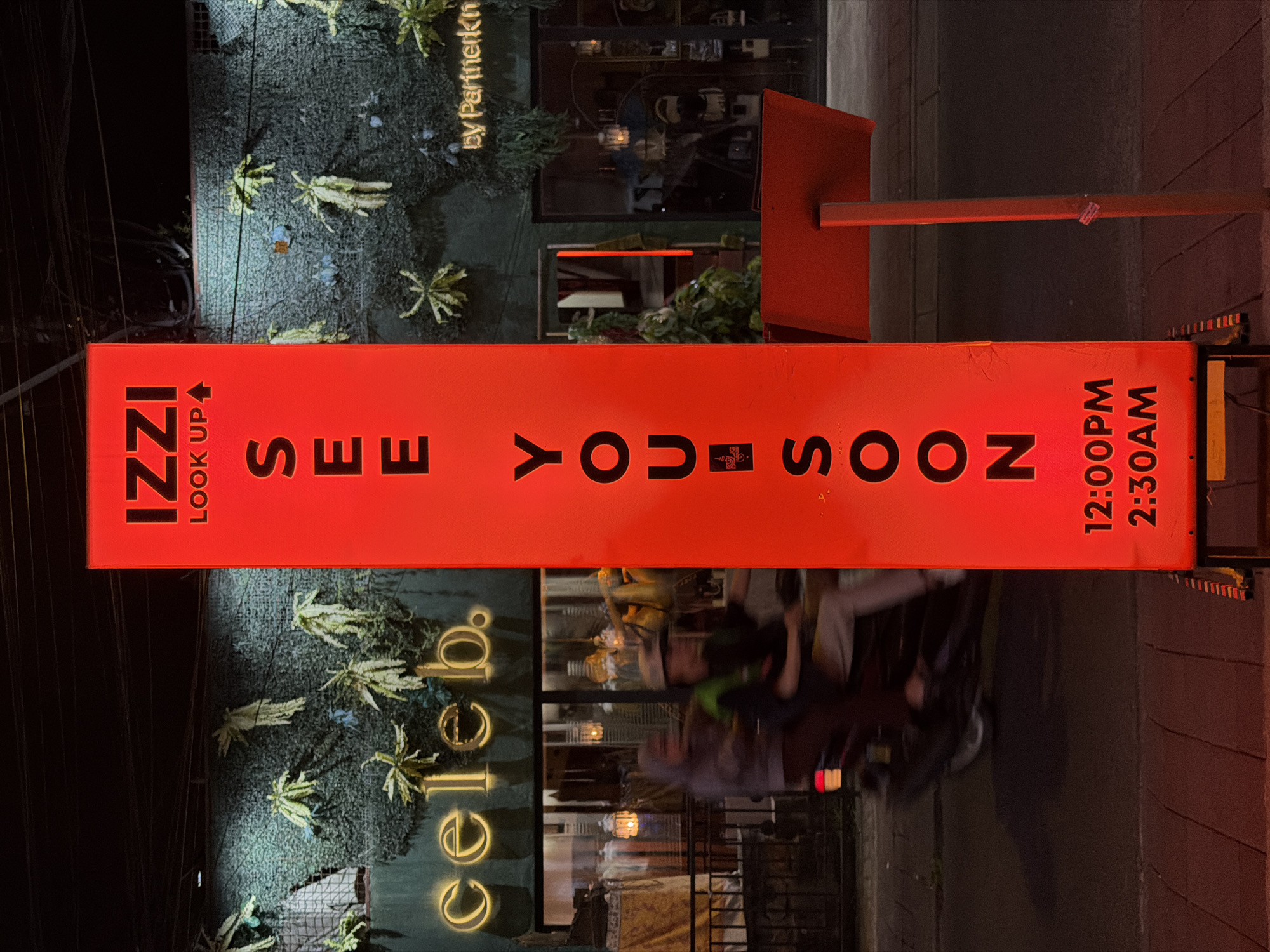 The image depicts an illuminated red sign that reads "IZZI LOOK UP SEE YOU SOON" with operating hours "12:00PM 2:30AM." The sign is located in front of a business named "Celeb." The business is "by PartnerKn," and the image was taken in North Kuta, Indonesia. A motorcycle is blurred in the reflection.