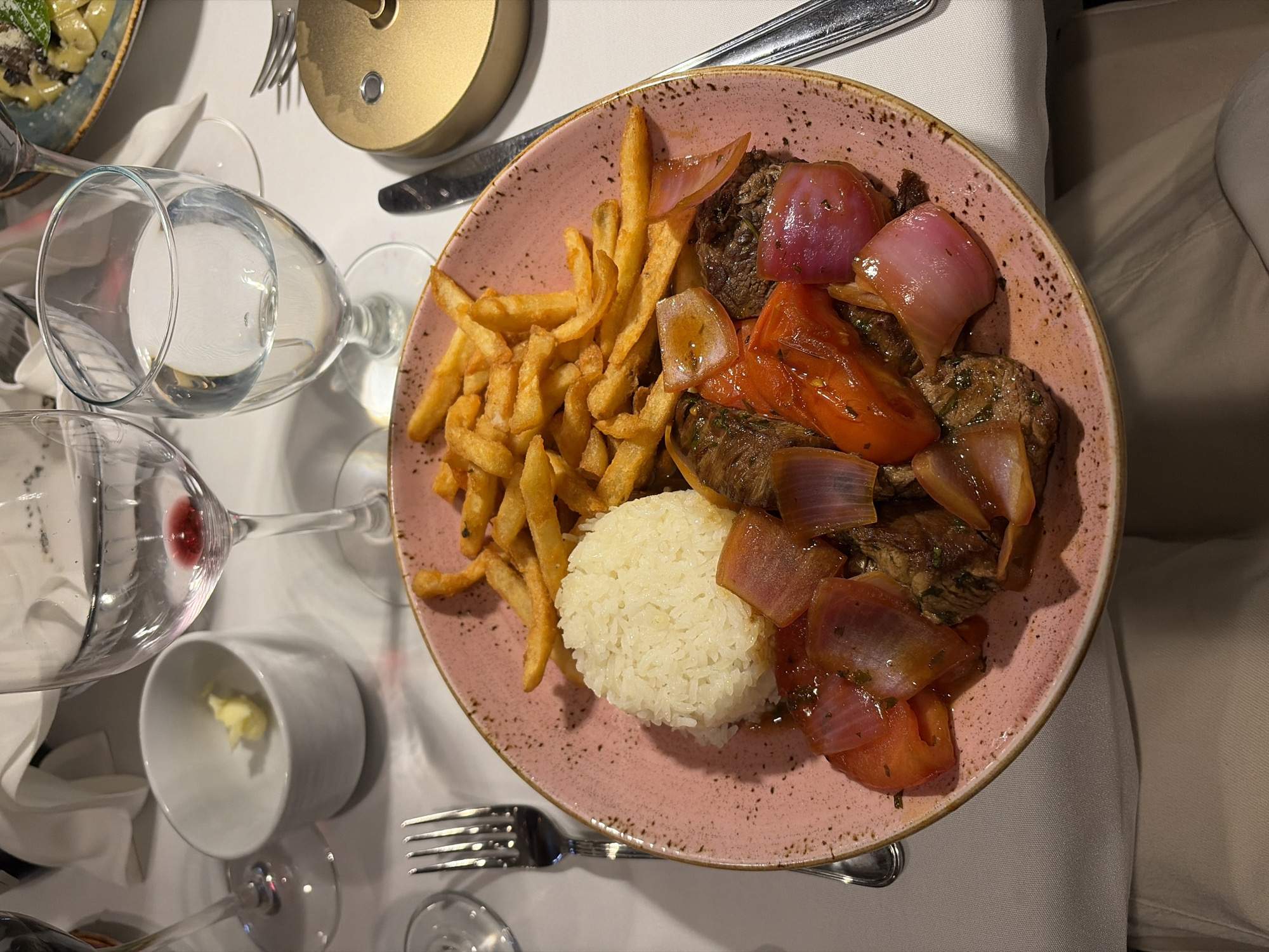 The image depicts a plate of Lomo Saltado, a traditional Peruvian stir-fry, consisting of marinated strips of sirloin, onions, tomatoes, French fries, and rice. The plate is pink and speckled, placed on a white tablecloth. Utensils are arranged around the plate, including a fork, knife, and spoons. Two wine glasses, one containing red wine, and a water glass are present, along with a small cup containing butter. The scene is likely set in a restaurant in Key Biscayne, United States of America.