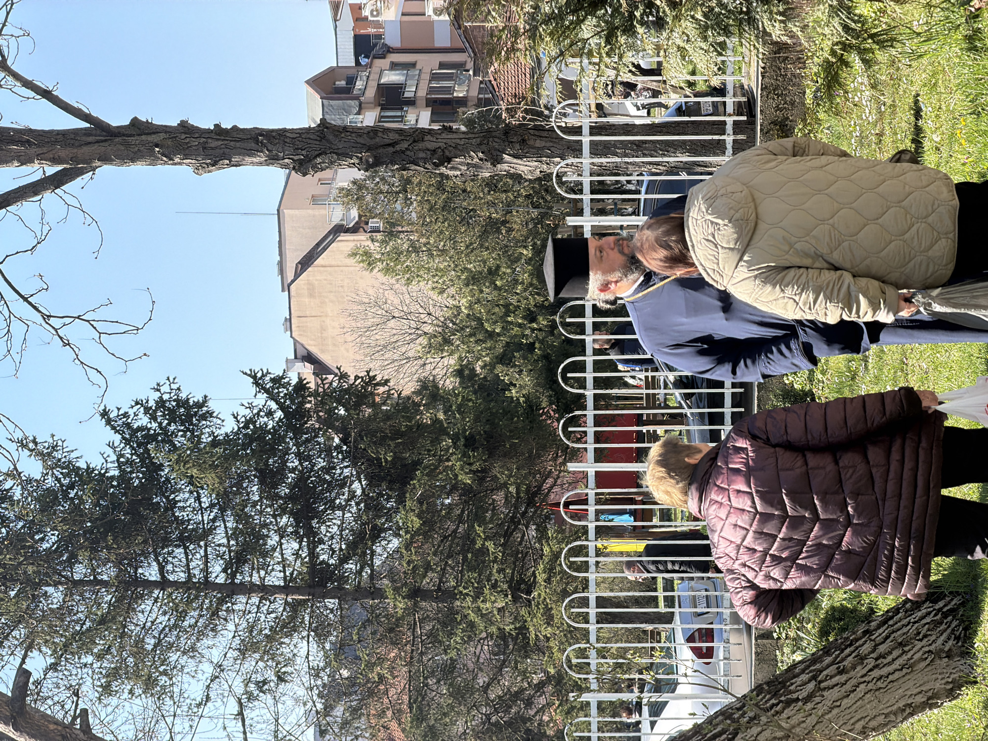 The image shows a scene in Sofia, Bulgaria. An Orthodox priest wearing a black hat and dark blue robe stands behind a white metal fence, speaking with two women. One woman wears a tan quilted jacket and the other wears a dark purple quilted jacket. They appear to be standing in a grassy area with trees. Behind the fence and the priest, there are parked cars, more trees, and apartment buildings. The sky is clear and blue.