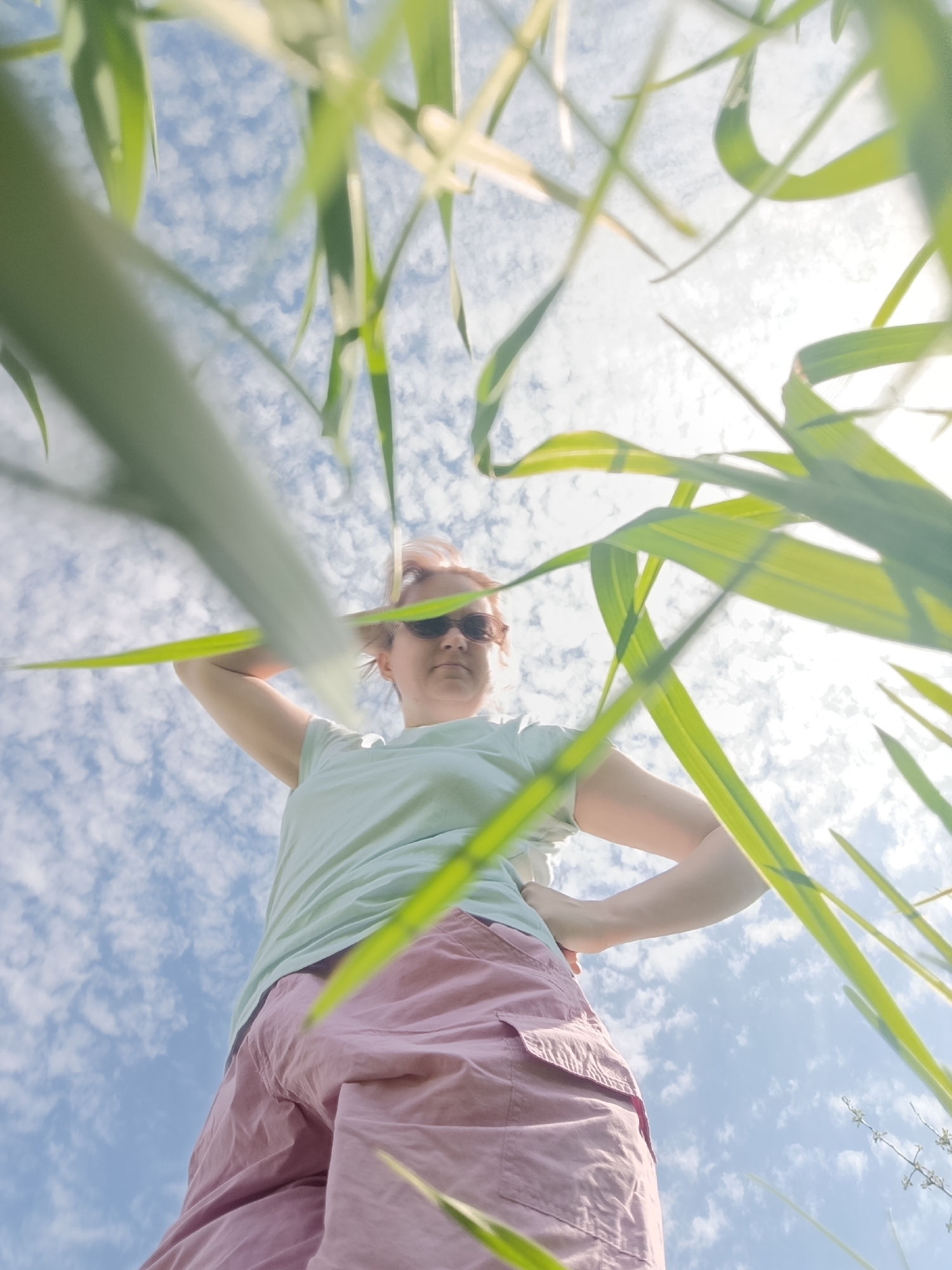 A low-angle shot shows a person with pink hair wearing sunglasses, a light green t-shirt, and pink shorts. They are standing with one hand behind their head and the other on their hip. The sky is visible between blades of green grass, with a mix of blue and white clouds and a bright light source. The photograph was taken in Dubovaya, Russia.