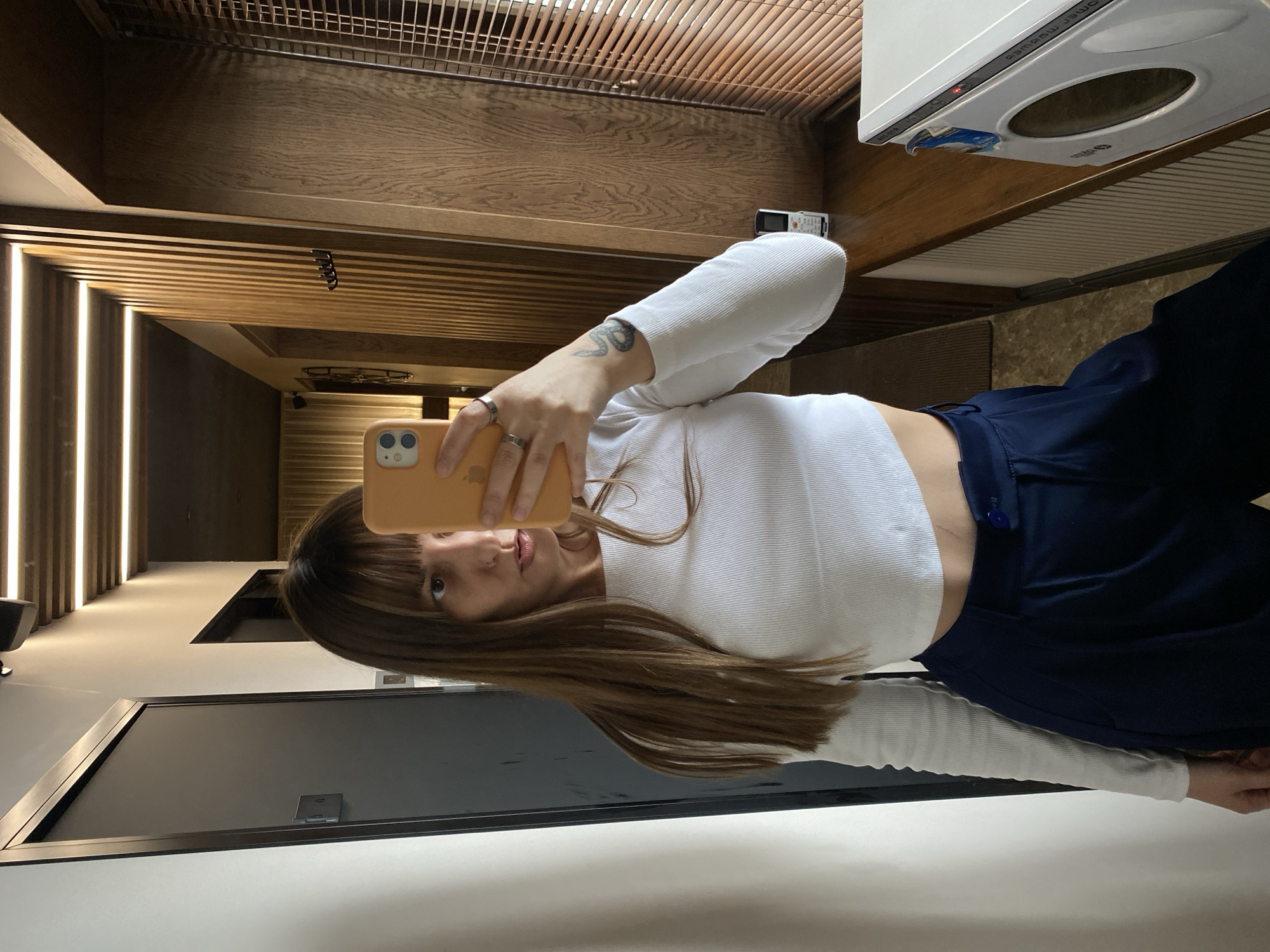 A woman is taking a selfie in what appears to be a modern apartment or hotel room. The woman is holding an orange smartphone, obscuring part of her face. She is wearing a white long-sleeved crop top and dark blue pants. She has long brown hair with bangs.
The background shows a hallway with vertical lighting and wooden ceiling details. A kitchen area is visible with a white appliance above her. A grey door is visible in the lower left corner.
The location of this image is unknown and there is no indication that this is in Votkinsk, Russia.
