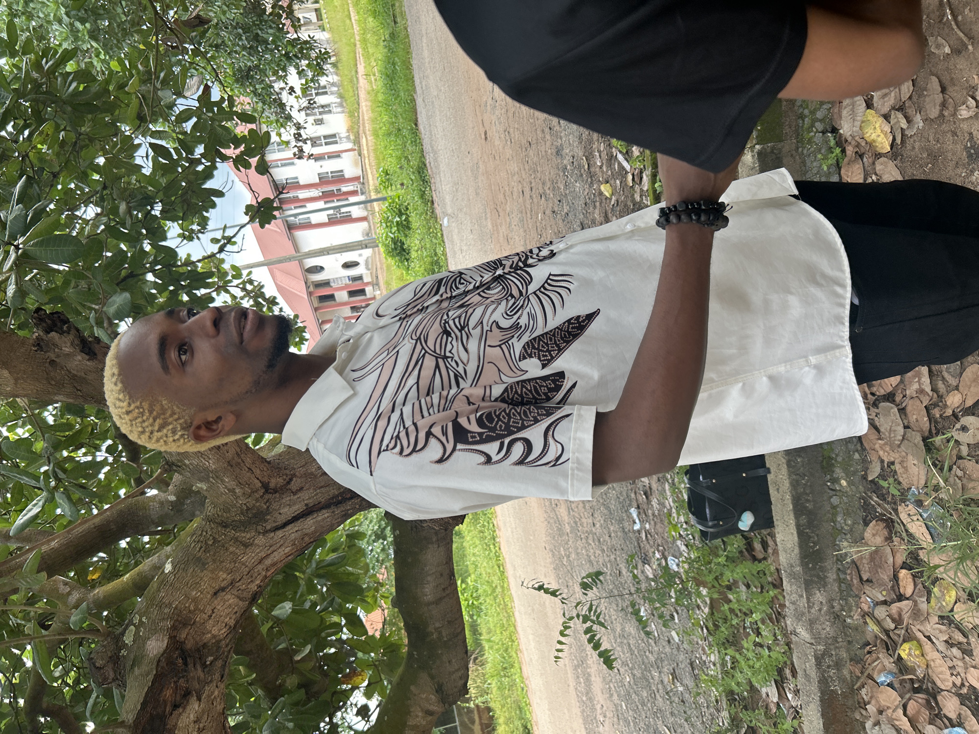A young man with blond dyed hair, wearing a white collared shirt with a black and pink abstract design, stands outdoors near a tree. He is looking upwards and to his left. On his left wrist, he wears a black beaded bracelet. He is also wearing black pants. Behind him, a dirt path runs alongside a grassy area and a building with red trim and white walls. The ground is covered with fallen leaves and some litter. The scene appears to be set in a rural or semi-rural area, consistent with the location context of Ihiagwa, Nigeria. The lighting suggests it is daytime, and the foliage indicates fair weather. There is no visible text in the image.
