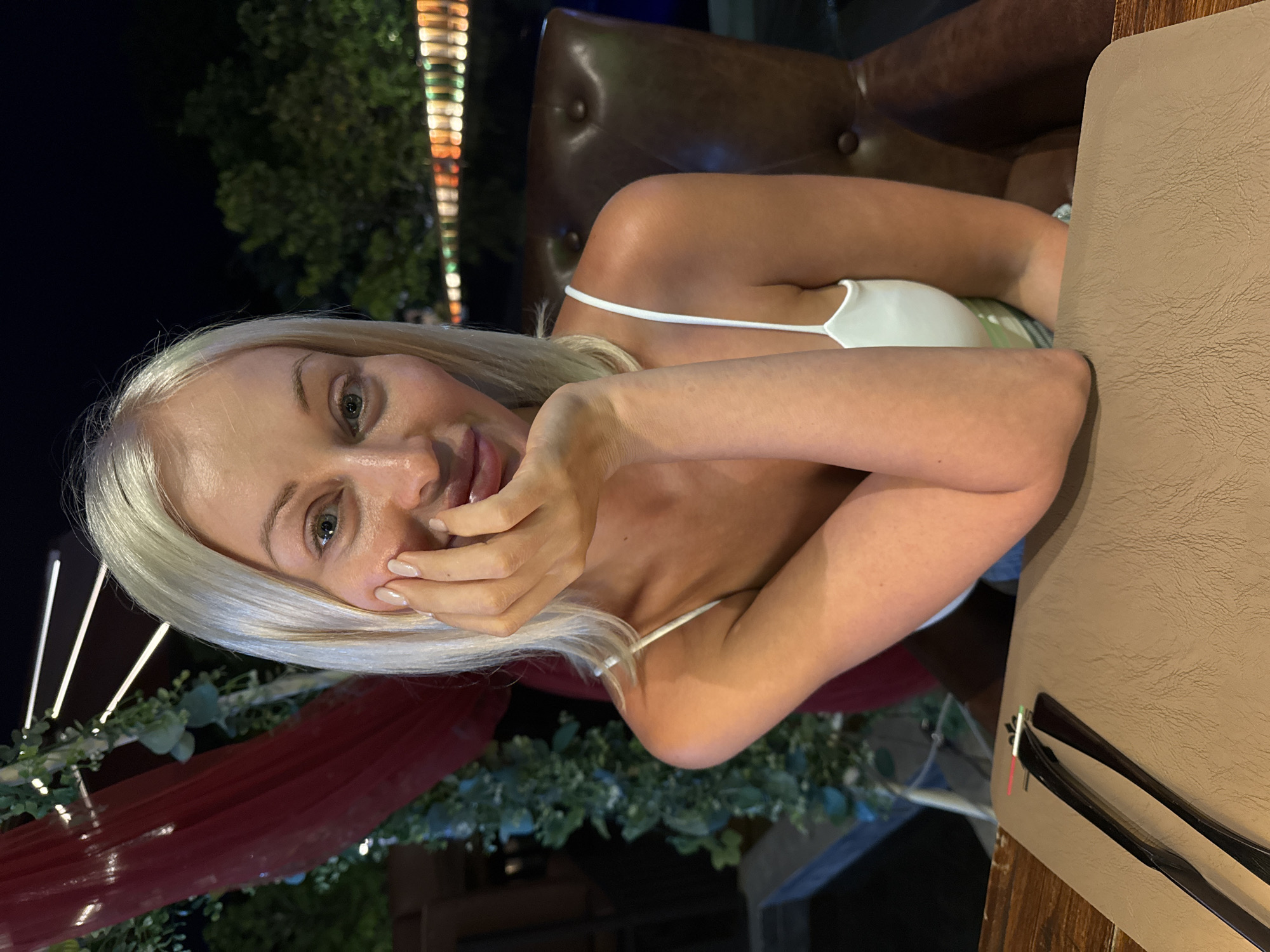 A woman with blonde hair wearing a white top is sitting at a table at an outdoor restaurant in Baan Chaweng, Thailand. The setting is at night, with decorative lights and greenery visible in the background. The woman has her hand near her face and appears to be smiling. Cutlery and a placemat are on the table in front of her. A leather chair is behind her.