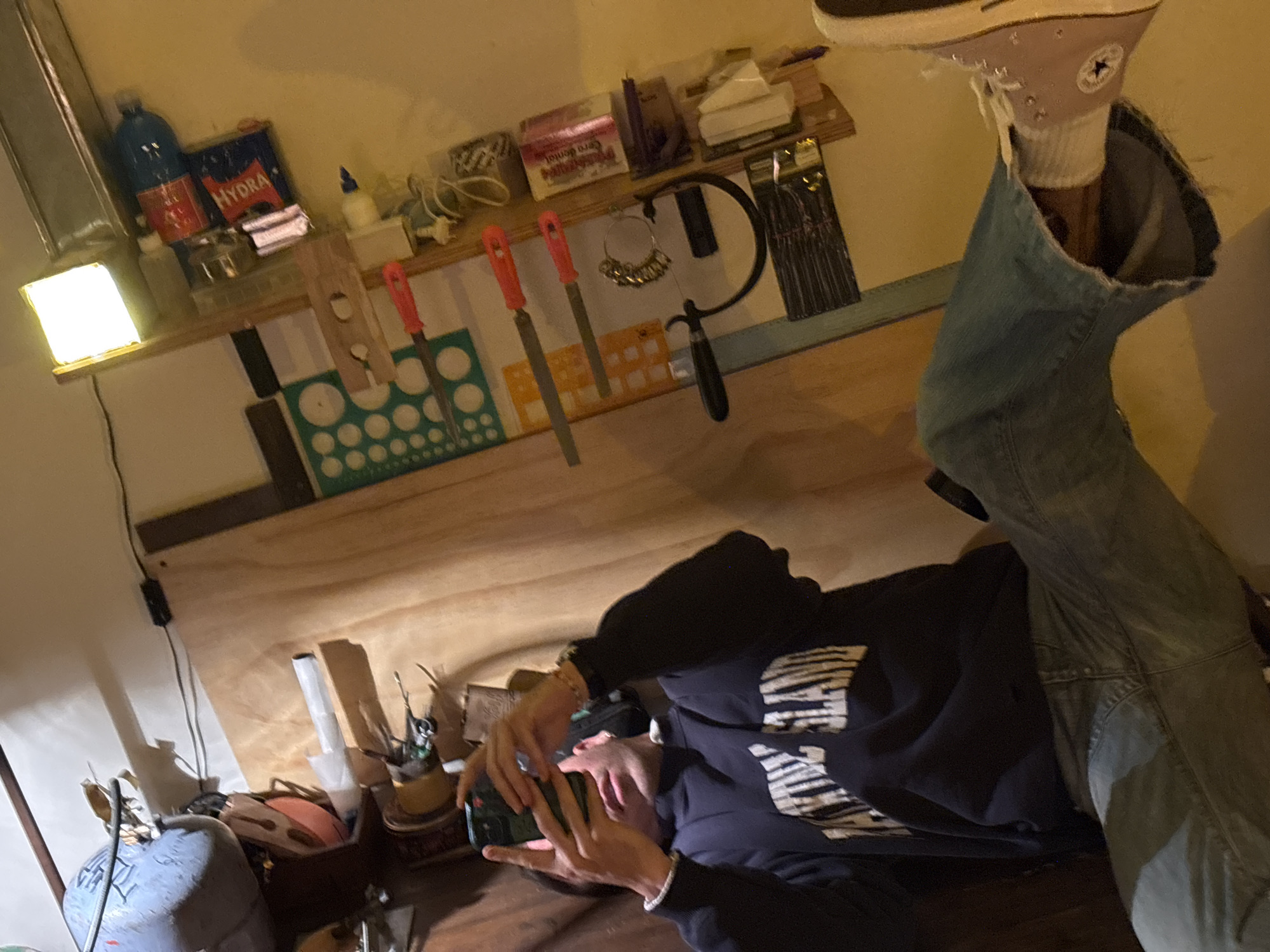 This overhead image depicts an individual lying on their back in a workshop or creative space in Buenos Aires, Argentina. The setting features a light wooden workbench and shelves, illuminated by warm artificial light, including a prominent bright, square LED light on the left.
The person is dressed casually in a dark hoodie with a light, patterned design, and light-wash blue jeans. One leg is raised, revealing a light-colored high-top Converse sneaker with the iconic "ALL STAR" logo and white socks. Both hands are holding a smartphone, suggesting they are actively using the device. Their posture indicates a moment of relaxation or a break from work.
The surrounding environment is rich with tools and supplies. On a shelf above, a blue gas cylinder and a box labeled "HYDRA" are visible. Other items include a box with "CERADENTAL" text, several red-handled files hanging from hooks, and a jeweler's saw frame holding a collection of rings or sizing gauges. A template with circular holes and a package of thin, dark items are also present. On the workbench surface, a gray gas tank is situated near various small containers holding tools like brushes, indicating activities such as jewelry making or fine crafting. The lighting creates a cozy, informal atmosphere.