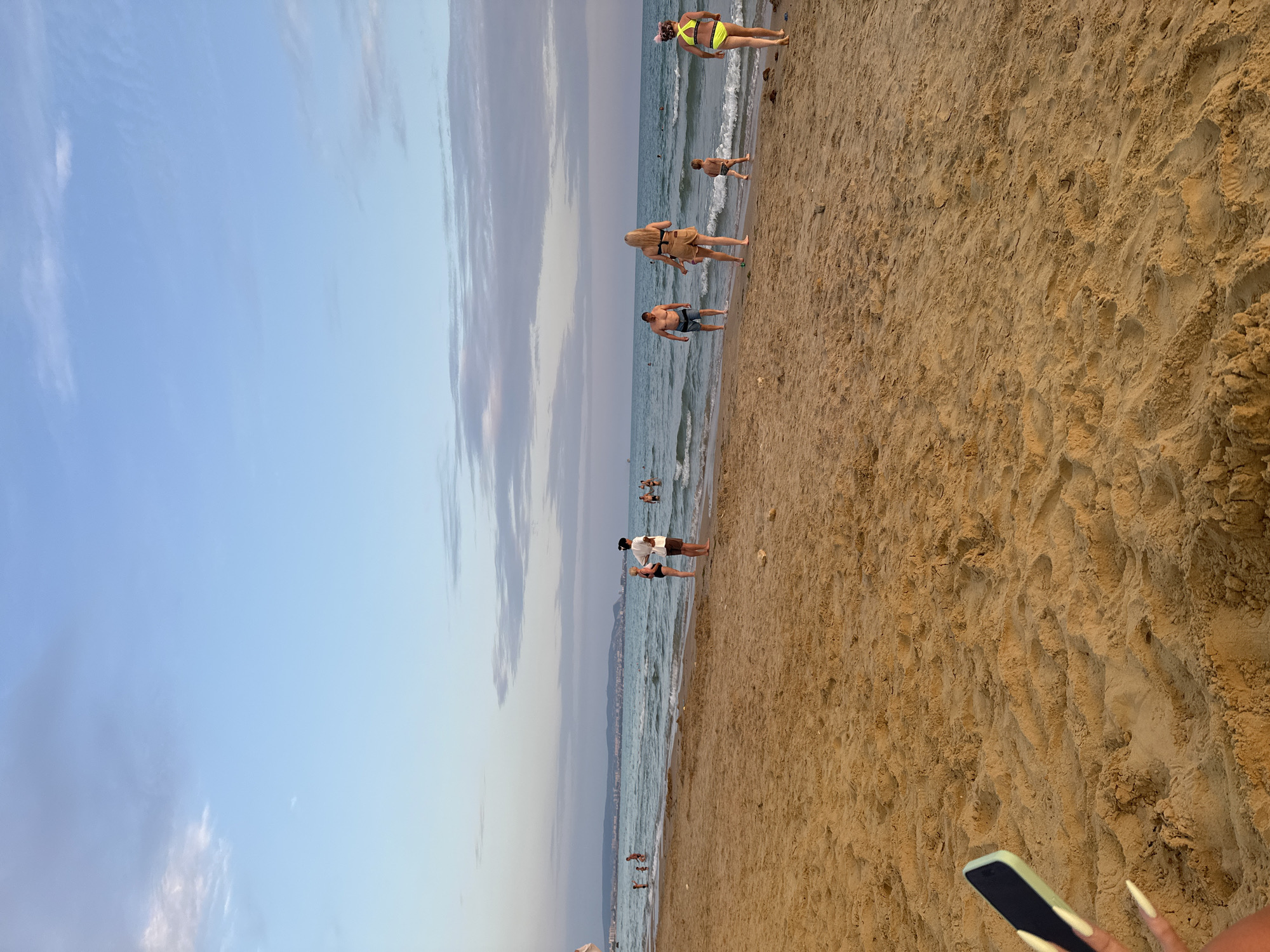The image depicts a sandy beach with people enjoying the water and shoreline. In the foreground, a portion of a smartphone is visible, held by a person whose hand with manicured nails is partially in frame. The beach is a light brown color with visible footprints and ripples in the sand.
The middle ground features several individuals walking along the shoreline and in the shallow water. A man in blue shorts walks towards the water. A woman in a bikini stands facing away from the camera, with a child in swim trunks next to her. Another woman in a bright yellow bikini walks away from the viewer, her back to the camera. Several other people are scattered further down the beach, some wading in the sea.
The background shows the calm sea with gentle waves, and a distant shoreline with buildings and a hazy mountain range under a pale blue sky with scattered clouds. The overall scene is peaceful, suggesting a relaxed atmosphere on a warm day. The location is identified as Vityazevo, Russia.