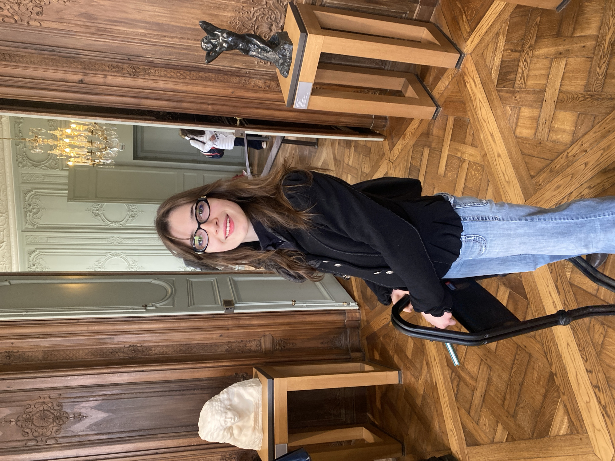 The image depicts an interior scene within the Musée Rodin, located in Paris, France. A young woman with long brown hair and black-framed glasses, dressed in a black jacket, dark turtleneck, and light blue jeans, stands front and center, smiling directly at the viewer. She is holding onto a black, portable folding stool. The floor is composed of polished light brown parquet laid in an intricate geometric pattern. The walls in the immediate foreground are paneled in dark, ornately carved wood. To the woman's left, a white marble bust, resembling "The Imploring Woman" by Camille Claudel, is displayed on a light wooden pedestal with a dark top surface. To her right, a dark bronze sculpture, resembling "Meditation" by Auguste Rodin, is presented on an identical pedestal with an information plaque. A light-colored doorway in the background opens into an adjacent room, revealing pale green walls adorned with elaborate white stucco molding and a prominent golden chandelier. A second individual, wearing a white coat and a dark backpack, is partially visible walking away through this doorway.