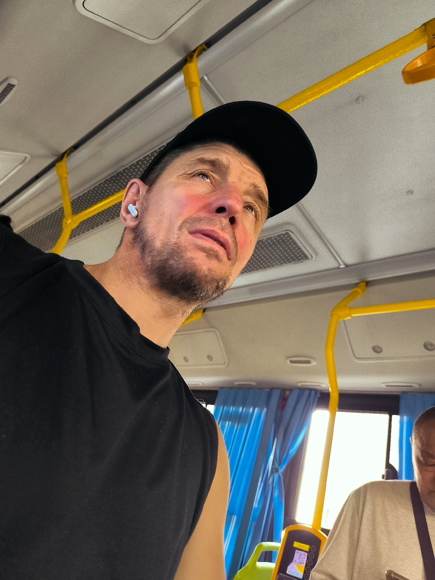 A man wearing a black t-shirt and a black baseball cap is inside a bus, looking upwards. He has a light beard and is wearing a white earbud in his left ear. The bus interior features yellow handrails, blue curtains, and a bright window. Another person, partially visible, wearing a light-colored t-shirt and a dark strap, is seated to the right of the frame. A yellow ticket dispenser with a screen displaying information is mounted on a green pole. The overall scene appears to be during daylight. The location is identified as the city administration of Qarağandı city, Kazakhstan.