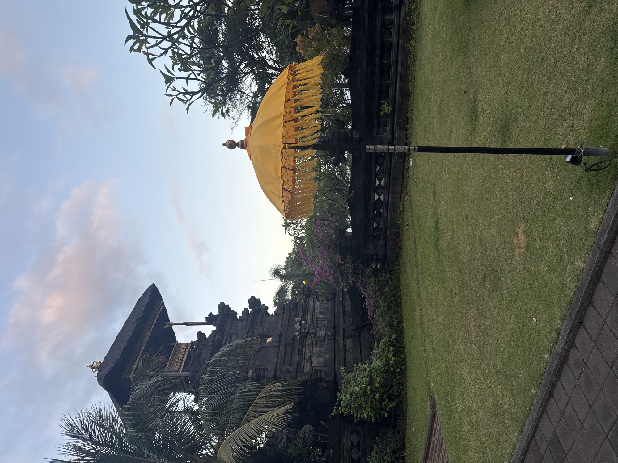 This image depicts a serene outdoor scene in Dawan, Indonesia, likely at a cultural site or temple complex. In the foreground, a vibrant yellow umbrella with decorative tassels stands on a black pole, positioned on a well-maintained green lawn. The lawn is bordered by a dark, stone-paved walkway on the left.
Behind the lawn, a traditional Balinese structure, possibly a temple pavilion, is visible. This structure is built from dark stone with intricate carvings and features a thatched roof topped with a golden ornament. Lush green foliage and purple flowering plants surround the base of the structure and line a decorative stone wall.
The background shows a clear sky with hints of pink and white clouds, suggesting either sunrise or sunset. Tall trees with dense canopies are also present, contributing to the tranquil atmosphere of the location. No people are present in the image. The overall impression is one of peace and cultural significance.