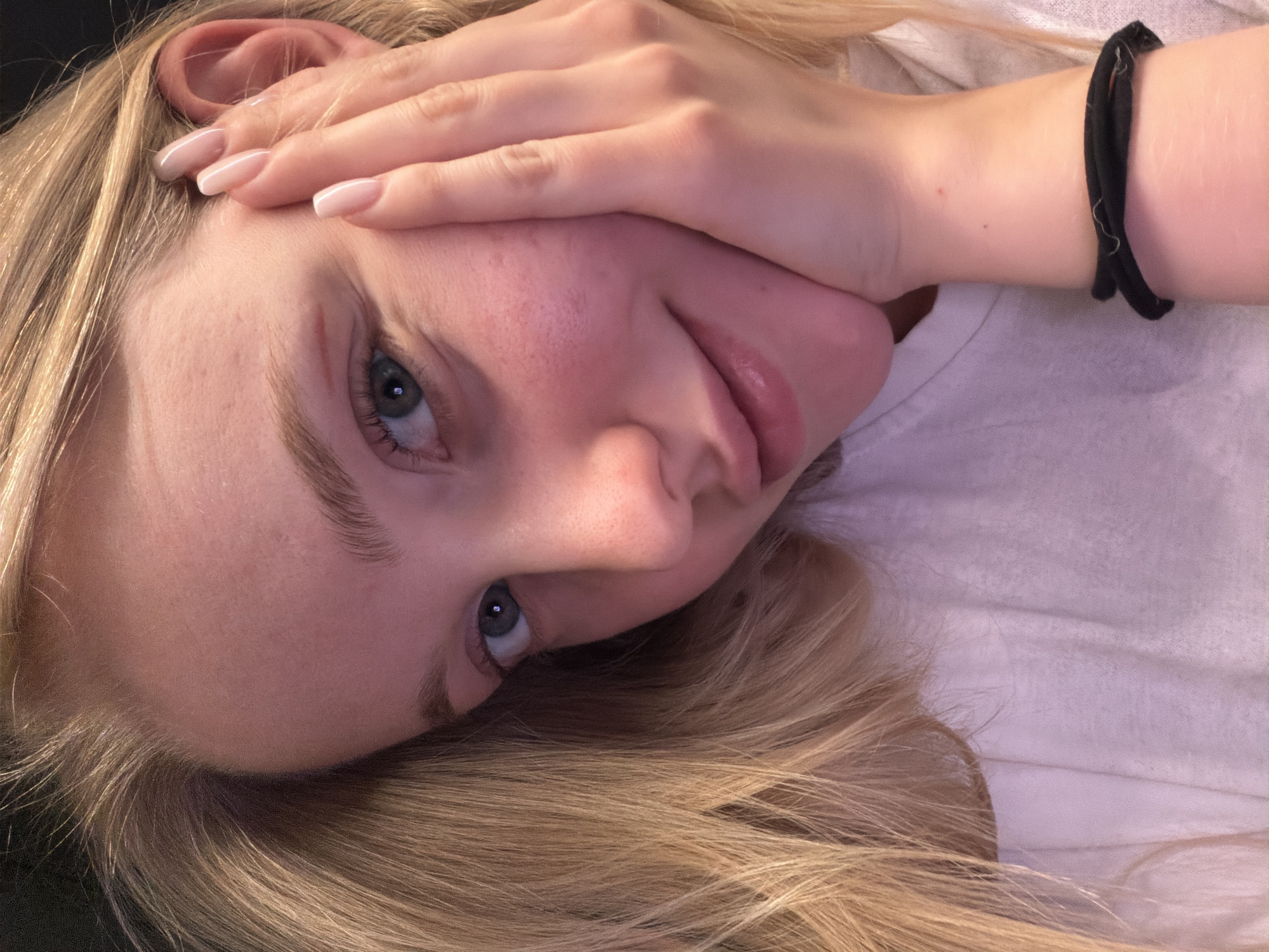 A blonde woman is lying down, looking up at the camera. Her right hand is on her forehead, obscuring part of her face. She has blue eyes and fair skin. She is wearing a light-colored shirt and a black elastic wristband. The background is out of focus. The location and city are unknown.
