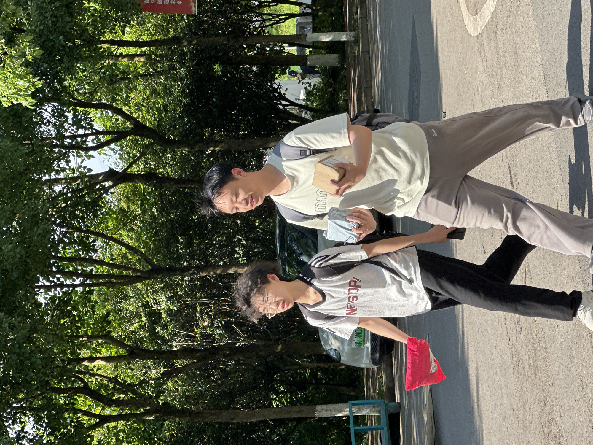 Two young men are captured in a vertical, full-body shot, walking outdoors on a sunny day in Jiangjin, China. The scene takes place on a paved surface, likely a street or pathway, bordered by lush green trees that cast dappled sunlight and distinct shadows.
The man in the foreground, on the left, has dark, curly hair and wears glasses. He is dressed in a light-colored t-shirt with grey sleeves and black stripes, bearing the red text "NFSUP!". He wears black pants and white sneakers, and carries a bright red bag in his left hand. A dark backpack is visible on his shoulders. He looks directly towards the viewer.
Behind him, slightly to the right, stands another man with dark hair, wearing a light-colored t-shirt, light grey sweatpants, and white sneakers. He has a dark backpack and holds a small, brown cardboard box in his right hand and a light blue/grey item, possibly a cloth or mask, in his left. He also appears to be looking towards the viewer with a slight smile.
In the background, a dark-colored car is partially visible behind the men, displaying a Chinese license plate that partially reads "A-AMO". The ground is a grey paved material with some white markings. The abundant green foliage indicates a vibrant natural setting, possibly a park or a tree-lined residential area. The bright sunshine and sharp shadows suggest it is daytime, likely morning or late afternoon. A partially obscured red sign with Chinese characters is visible among the trees in the upper left background, and a blue metal barrier is near the ground on the left.