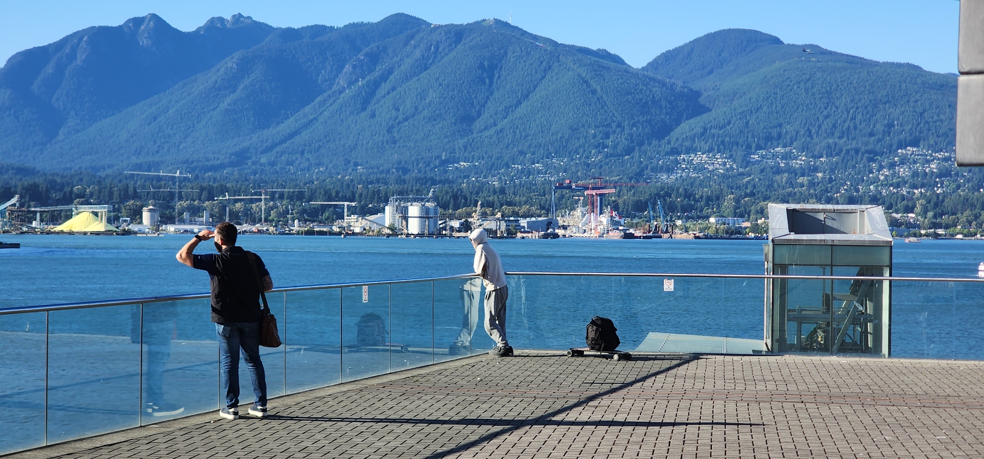 Two individuals are observed on a waterfront promenade overlooking a body of water and a distant mountainous cityscape. The person on the left, facing away from the camera, wears a black polo shirt, blue jeans, and sneakers, and is holding their right hand up to shield their eyes while looking towards the water. They are also carrying a brown satchel over their left shoulder. The second individual, positioned further to the right and closer to the water, is wearing a hooded light-colored sweatshirt and grey pants. They are leaning on a glass railing, facing towards the water. Adjacent to this person is a black backpack placed on a skateboard. The environment features a tiled walkway, a glass railing, and a modern glass structure to the right. In the background, the water is calm, and across it, industrial buildings with cranes are visible against a backdrop of densely forested mountains dotted with houses. The sky is clear and blue.
