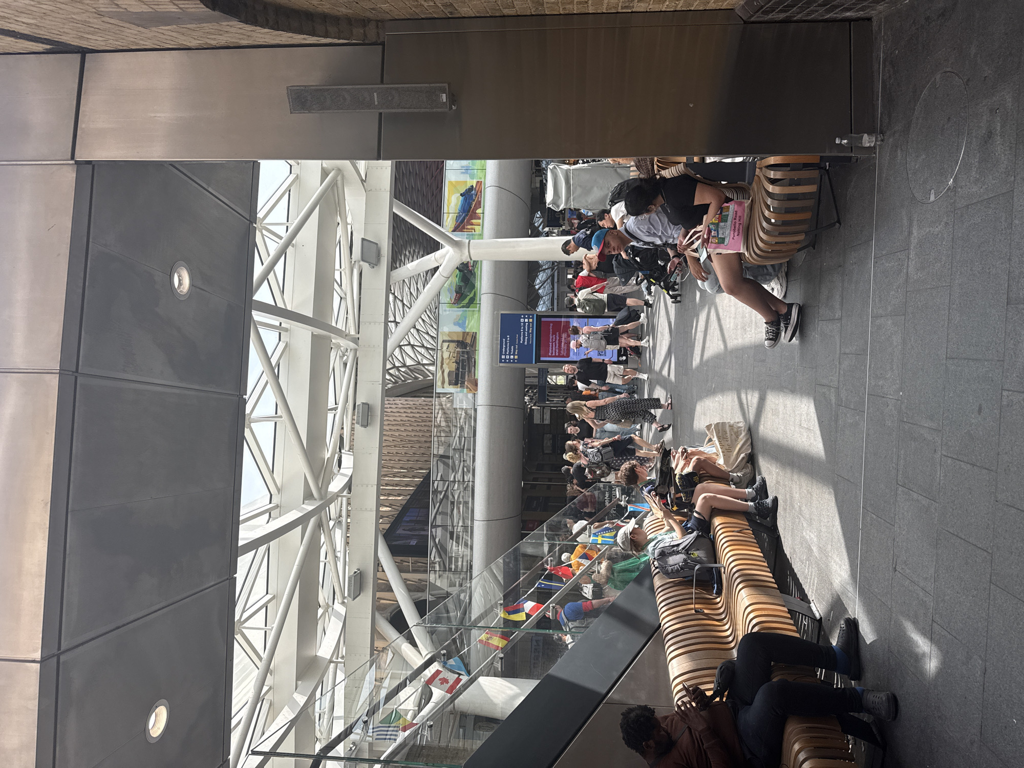 A horizontally-oriented image shows a public area with a modern architectural design. People are seated on a series of curved wooden benches that stretch into the distance. Some are using electronic devices, while others appear to be waiting.
Flags from various nations are displayed behind a glass railing that parallels the benches. The building features a high ceiling supported by a network of white, steel beams and glass panels, allowing natural light to illuminate the space.
The scene implies an urban area within Unknown, Unknown.