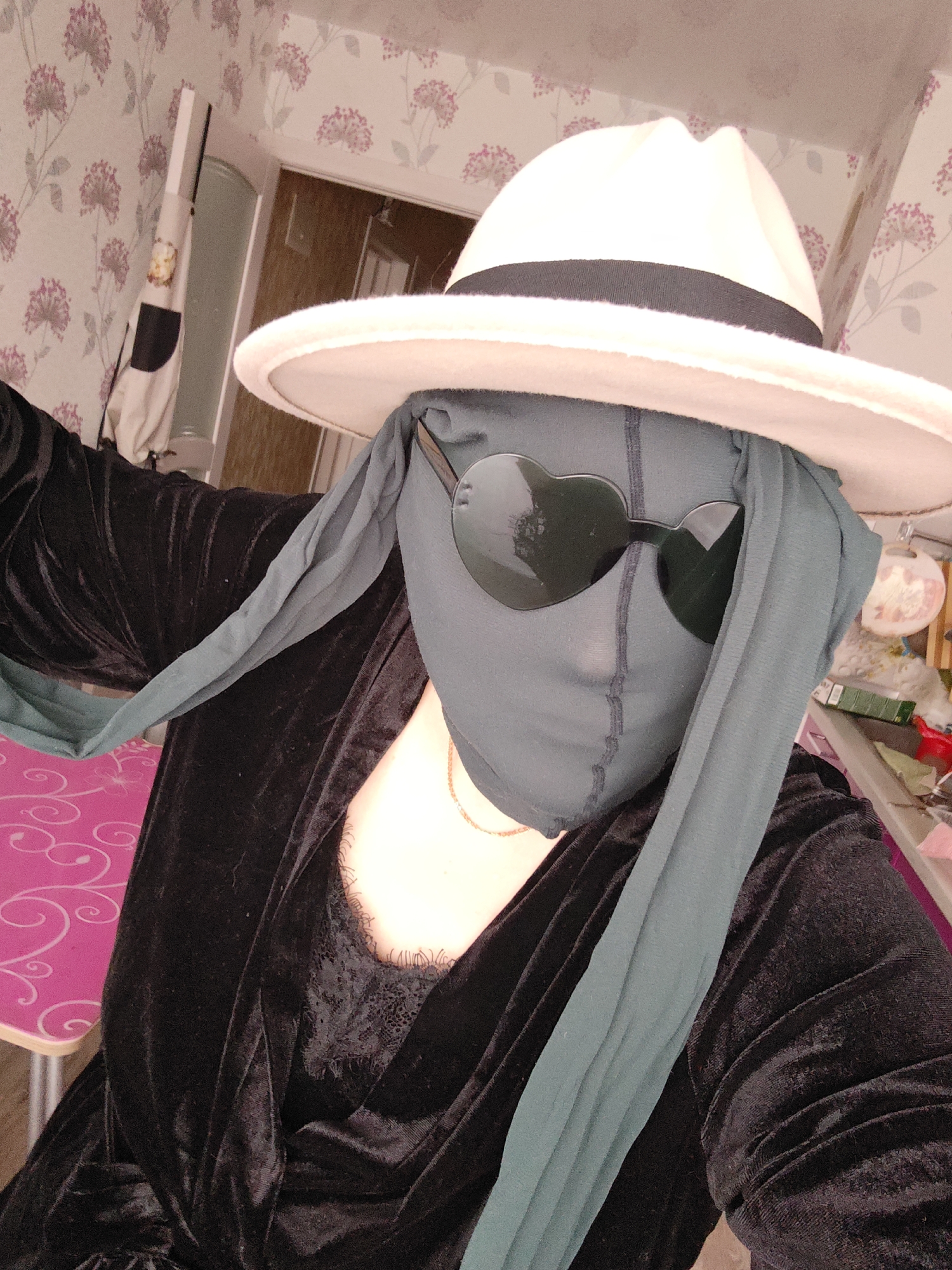 A person is posing indoors. They are wearing a white fedora with a black band, heart-shaped sunglasses, a grey face covering, and a black velvet robe with lace detailing at the chest. A gold necklace is visible at the neck. The background includes floral wallpaper, a doorway, and items on a shelf. There is a pink table or surface visible on the left. The location appears to be a cluttered interior space. There is no indication this image was taken in the city of Markovskii, Russia.