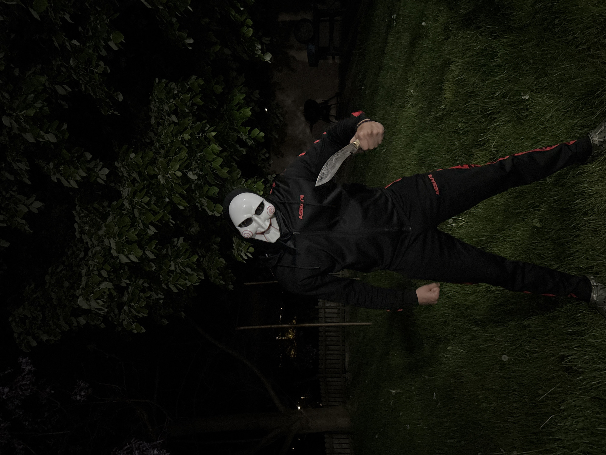 A person wearing a "Jigsaw" mask and dark clothing is standing on a lawn at night. The person is holding a large knife. The location appears to be a backyard or park with trees and grass. The location in Goritsa, Bulgaria is not definitively identifiable in the image, as identifying the location with certainty requires more information beyond what is present in the image.