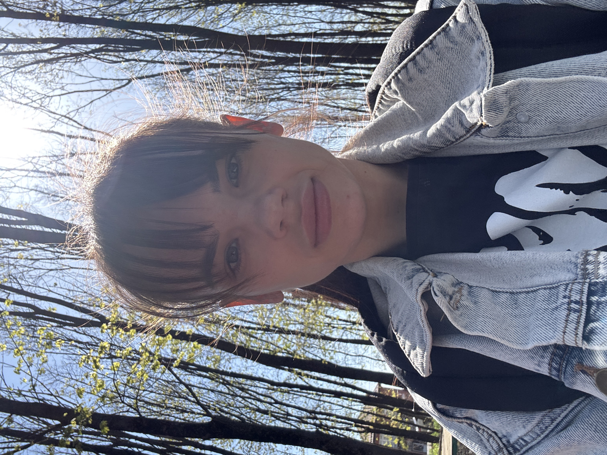 A bright, rotated outdoor selfie captures a young woman looking directly at the camera with a neutral to slightly smiling expression. The image is oriented with her head on the left and her shoulders on the right. She has dark hair with bangs, and her face is illuminated by natural daylight.
She is wearing a light blue, faded denim jacket over a dark t-shirt, which features a prominent white, abstract graphic that resembles a stylized skull or a similar artistic design.
The background consists of a clear blue sky, partially obscured by a dense network of bare tree branches. Some of these branches show very early signs of new, light green leaves or buds, indicating the season is early spring. Intense sunlight is visible as a strong glare on the upper left side of the image, backlighting her hair and creating a bright halo effect.
The scene depicts a casual outdoor moment, likely in a park or a natural setting within Rostov-on-Don, Russia, during a sunny day. No readable text is visible, only the abstract graphic on her shirt.