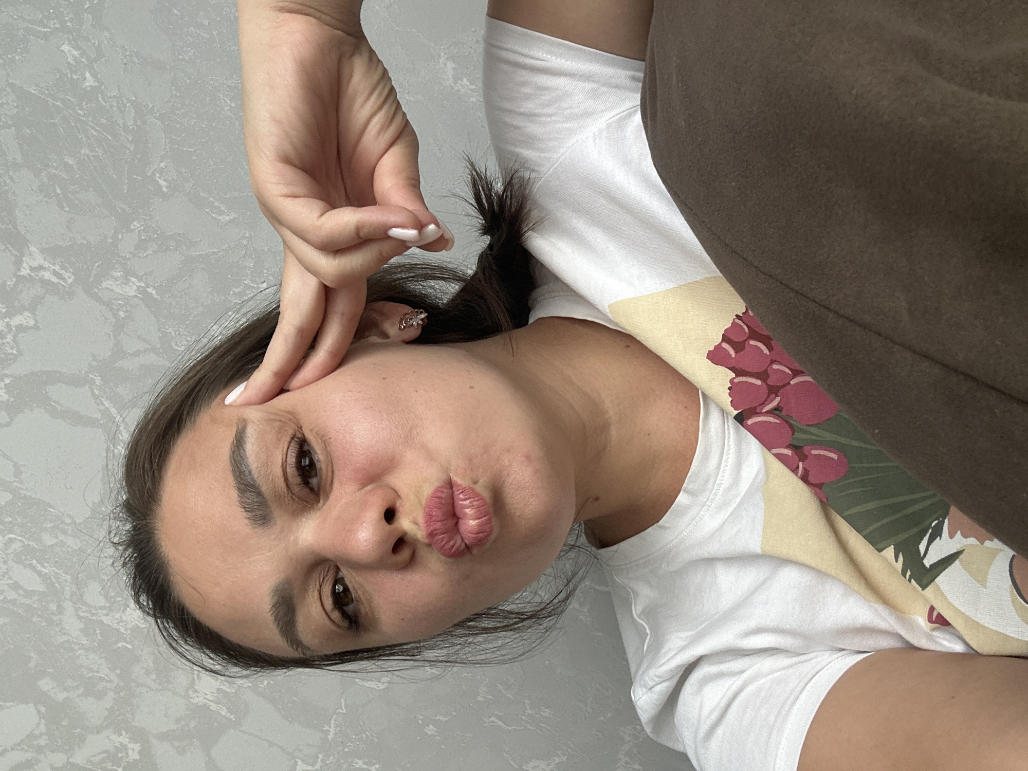 A young woman with dark hair and brown eyes is lying down, looking at the camera. She is wearing a white t-shirt with a floral design on it and brown pants. Her left hand is resting on her forehead, and her lips are pursed. The background is a textured gray wall. The image appears to be taken indoors. The location is unknown.