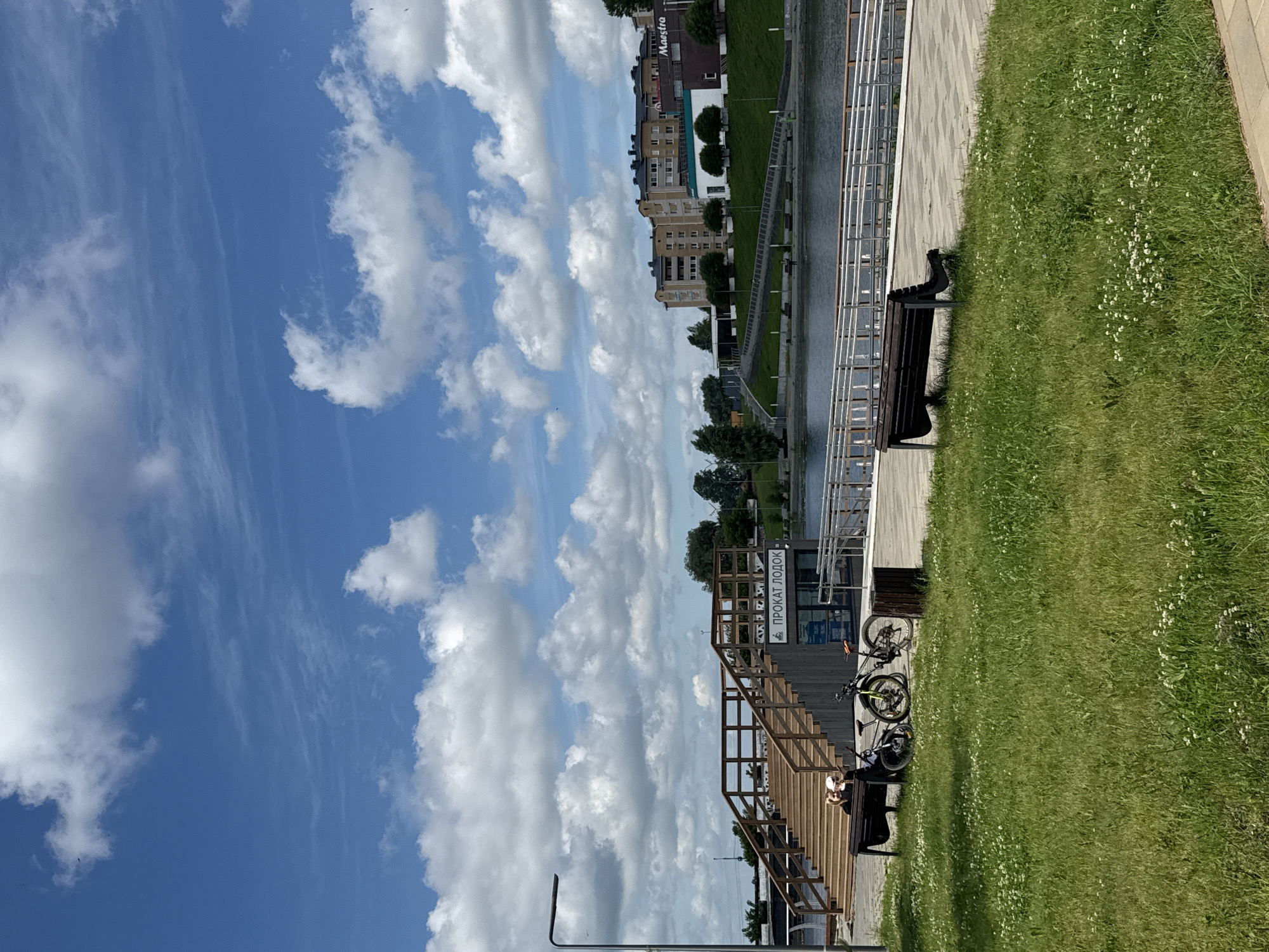 This is a photograph of a waterfront area in Votkinsk, Russia. In the foreground, a lush green lawn is dotted with small white wildflowers. A dark wooden park bench sits on a paved walkway in the lower right of the frame. In the middle ground, a wooden staircase leads up to a building with the sign "ПРОКАТ ЛОДОК" (Boat Rental) visible. Two bicycles are parked near the bench in front of the staircase. Two people are sitting on the bench at the base of the stairs, one leaning back and the other looking at a phone. A dark trash receptacle is next to the bicycles. Beyond the building, a body of water, likely a river or lake, is visible with a ramp and railing along its edge. In the background, across the water, are residential buildings and some trees. The sky above is a vibrant blue with scattered white cumulus clouds. A modern lamppost stands on the left side of the frame. The overall scene depicts a pleasant day at a recreational waterfront.