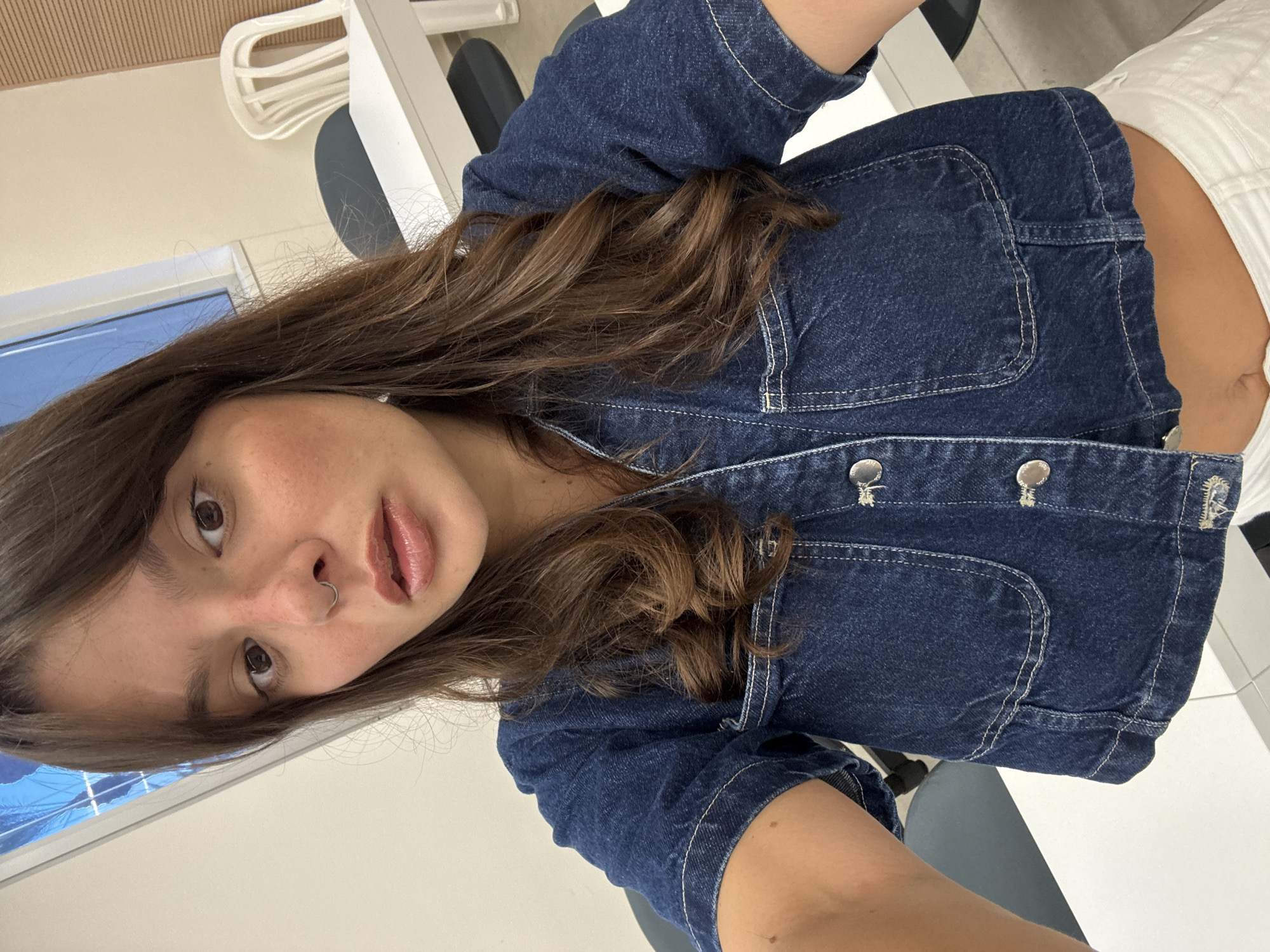 A woman with dark brown hair and a nose ring is taking a selfie in what appears to be a classroom or office setting. She is wearing a blue denim jacket over a white top, the latter exposing her midriff. The background shows white tables and chairs, and a window with a blue tint, possibly part of a building in Tel Aviv, Israel.