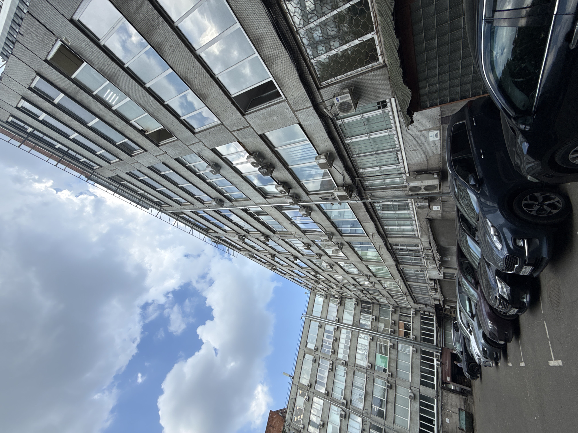 The image shows a narrow urban scene in Khimki, Russia, dominated by two gray, multi-story buildings facing each other. Their facades are characterized by repetitive rectangular windows, many reflecting the blue sky and white clouds. Numerous air conditioning units are attached to the exterior walls of the buildings. Parked cars line the street on the right side of the frame.