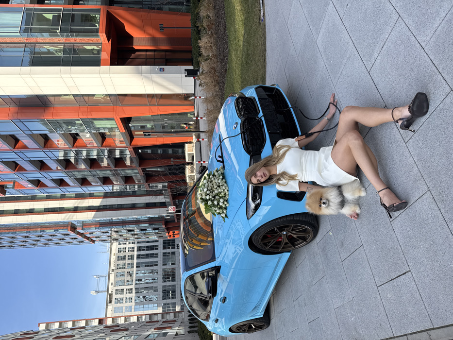 The image displays an outdoor scene featuring a woman, a dog, and a light blue BMW sedan against a backdrop of modern architecture. A light-skinned woman with long, light-colored hair, dressed in a short-sleeved white dress and black high-heeled sandals, is seated on a grey paved surface. Her legs are extended, and she leans on her left hand. She holds a black leash in her left hand, connected to a light brown Pomeranian dog seated to her left. A black cable extends from the front grille area of the BMW, resting on her right wrist. The car's hood is adorned with a large bouquet of white roses. The BMW features dark multi-spoke wheels and red brake calipers. The background consists of several modern, multi-story buildings with prominent orange and white facades and extensive glass windows. A patch of green grass is visible to the right of the car. The scene is brightly lit under a clear sky. The architectural style indicates the Żoliborz district in Warsaw, Poland, characteristic of the Żoliborz Artystyczny development.