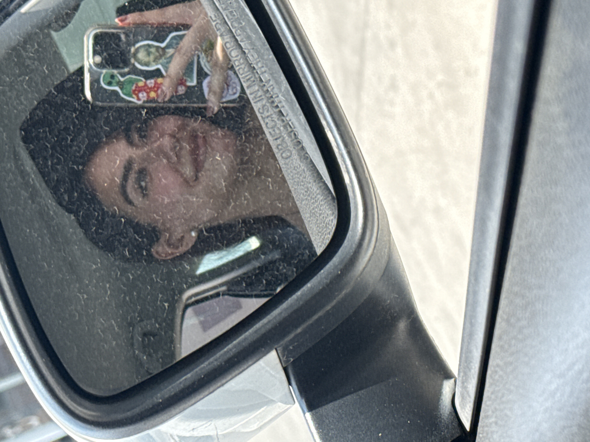 The image shows a reflection in a car's side mirror. The reflection appears to be of a young woman holding a mobile phone with a decorated case. The mirror is dusty. The surroundings are bright. The mirror has text on it that reads "OBJECTS IN MIRROR ARE CLOSER THAN THEY APPEAR". The city of Puebla, Mexico is not identifiable from the provided image.