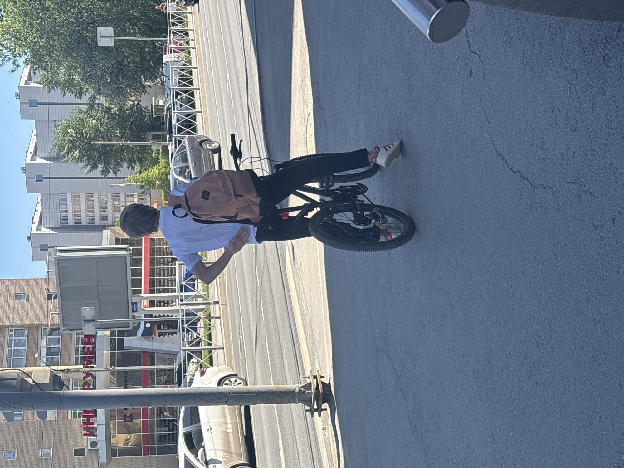 A young person rides a bicycle on a city street in Perm, Russia. The individual wears a light blue shirt, black pants, and light-colored shoes with red accents. A brown backpack is slung over their shoulder. They are biking next to a street with white dividing lines, bordered by a metal fence and a sidewalk. Cars are parked along the street, and buildings are visible in the background, including one with the word "Бумеранг" in large red letters. A traffic light pole is in the foreground. The environment suggests an urban setting on a clear, sunny day.