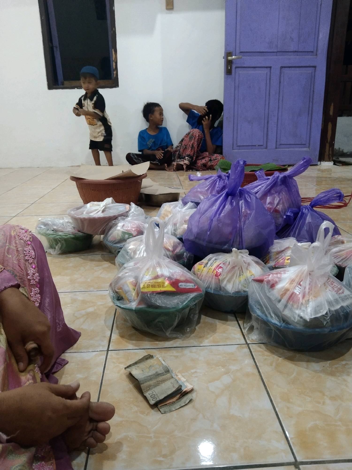 The image depicts a domestic scene in Buntalan, Indonesia, likely a home interior. The floor is tiled in a beige and brown pattern. In the foreground, the lower legs and feet of a person are visible, wearing a purple and pink patterned garment. Next to the person's feet, on the floor, are several Indonesian Rupiah banknotes, with denominations of 2000 and 5000 visible.
In the mid-ground, several plastic bags filled with what appear to be food items or provisions are arranged on the floor. Some are transparent, showing the contents, while others are opaque purple bags. These bags are placed in and around various bowls and baskets. A brown woven basket is prominent in the upper mid-ground.
In the background, three children are present. A young boy wearing a blue cap and a black and beige t-shirt stands near a window. To his right, another boy in a blue t-shirt sits on the floor. Further to the right, a third child, possibly an older child or young adult, is seated, with their hands near their head, and is wearing a patterned skirt. A large, closed purple door is visible in the far right background. The wall behind the children is white. The overall impression is one of preparation or distribution of food, possibly for an event or as aid.