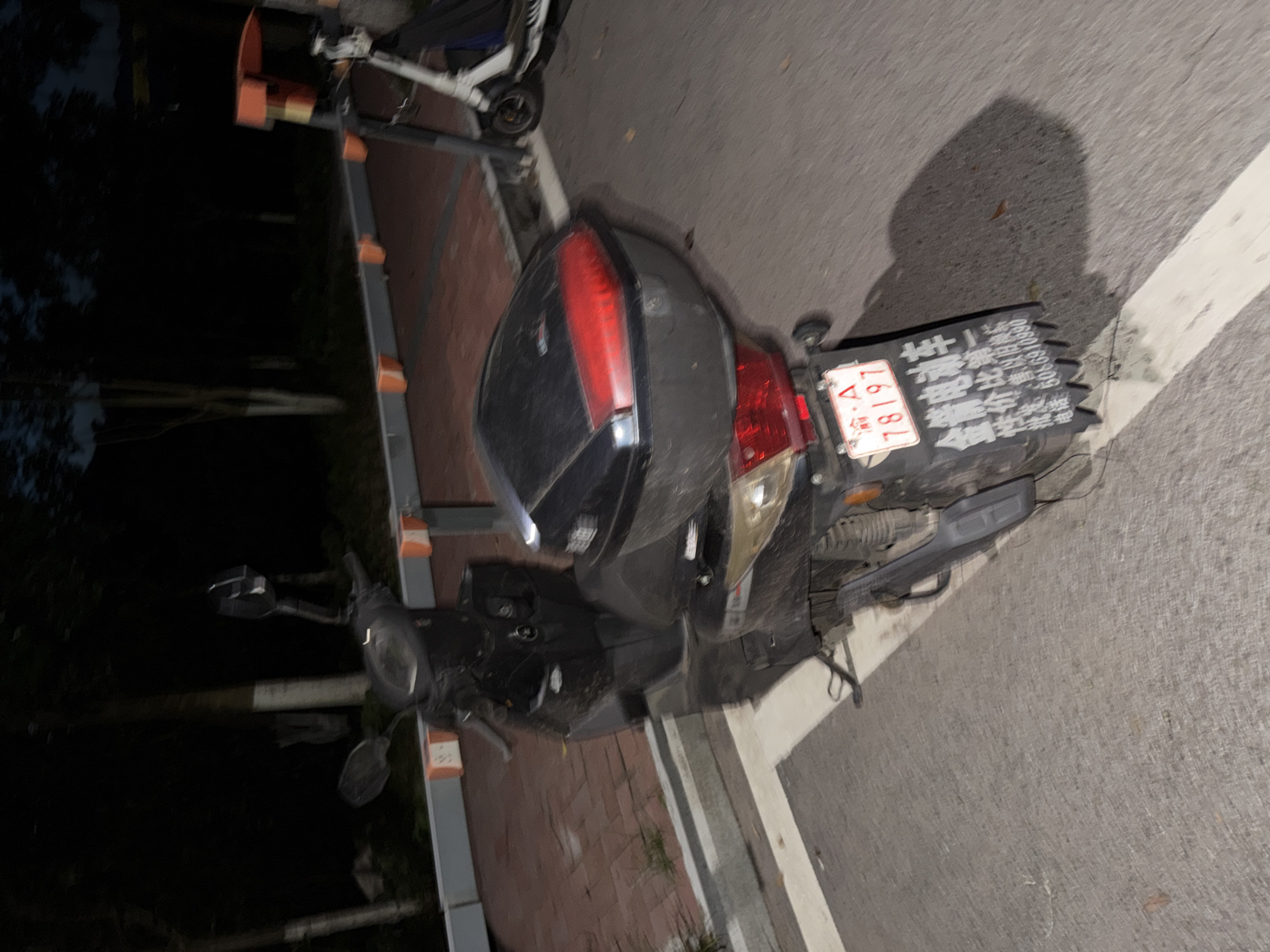 A black scooter is parked on an asphalt road in front of a brick walkway lined with metal barriers. The scooter has a red taillight and a black storage box on the back. A license plate with Chinese characters and the number "78197" is attached to the back of the scooter. A white electric scooter is parked behind the black scooter. The scene is dimly lit, with trees and dark shadows in the background, suggesting it is nighttime. The location is identified as Jiangjin, China.
