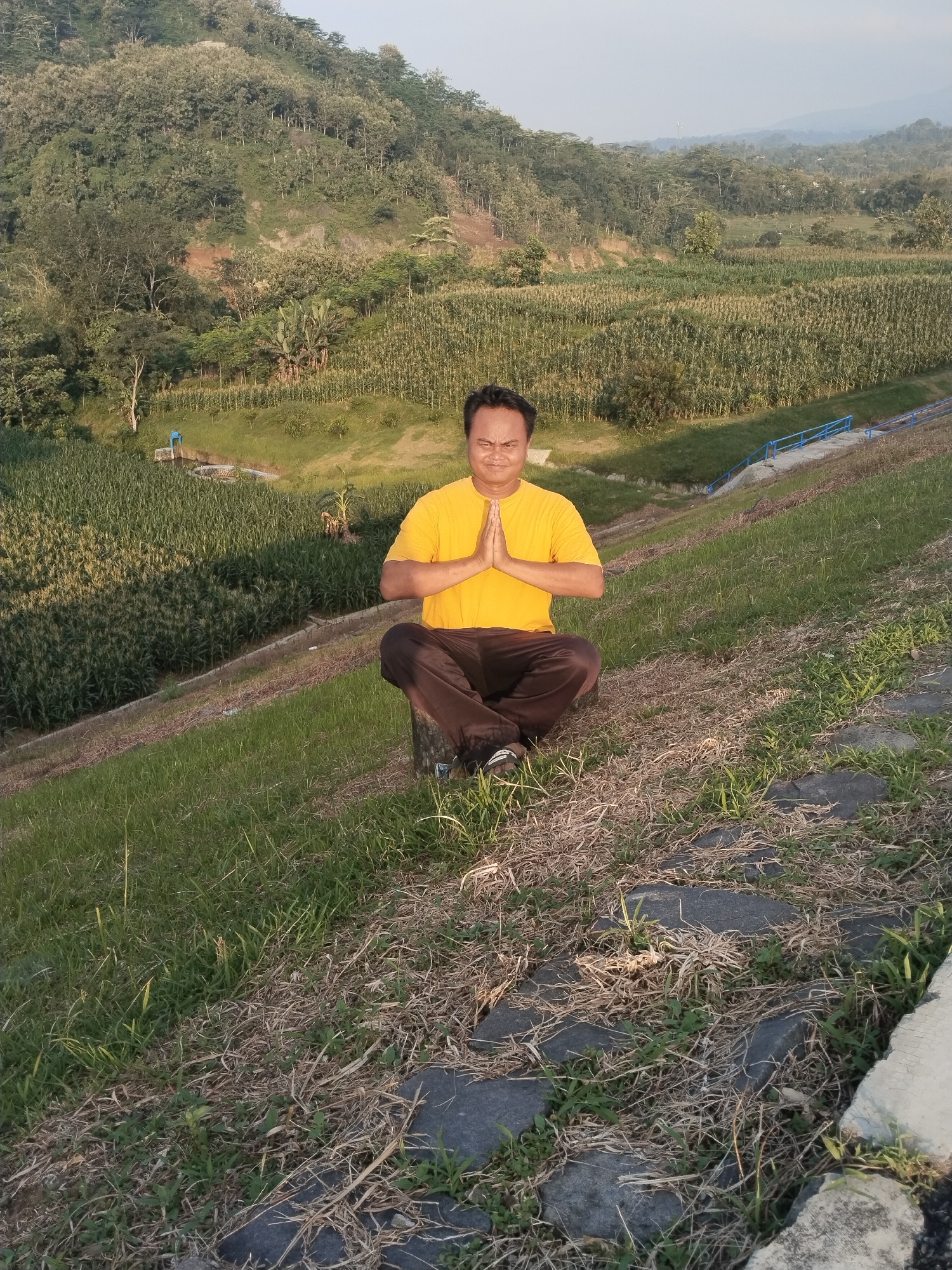 The image shows an individual seated in a meditative pose outdoors. The person, wearing a yellow t-shirt and brown pants, is cross-legged with hands pressed together in front of the chest. The setting appears to be a grassy slope with patches of dry vegetation, possibly a dam or embankment, with steps made of natural rock.
In the background, there's a field of what appears to be crops (perhaps corn) leading up to a forested hillside. A small structure and a blue railing are also visible in the background. The scene is set in daylight, the location being Jambeyan, Indonesia.