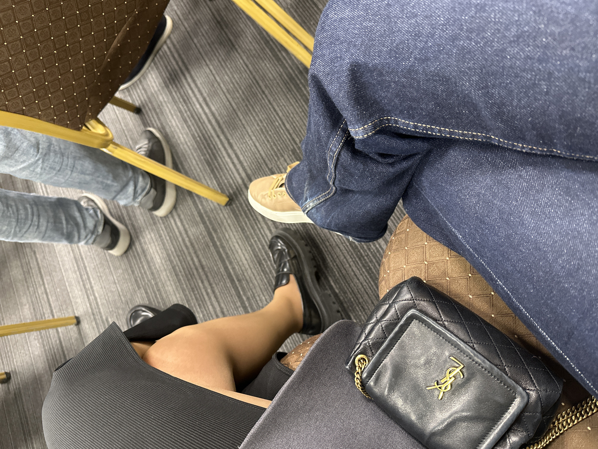 The image shows a view from above, focused on the legs of three seated individuals. One person wears blue jeans and tan shoes. Another person wears gray jeans and gray sneakers. The third person, possibly a woman, wears a black dress, black shoes, and carries a black YSL purse. All are seated on chairs with a gold-colored frame. The floor appears to be a neutral gray tone. The location is not discernible from the image content. Armenia or Yerevan is not indicated in the image.