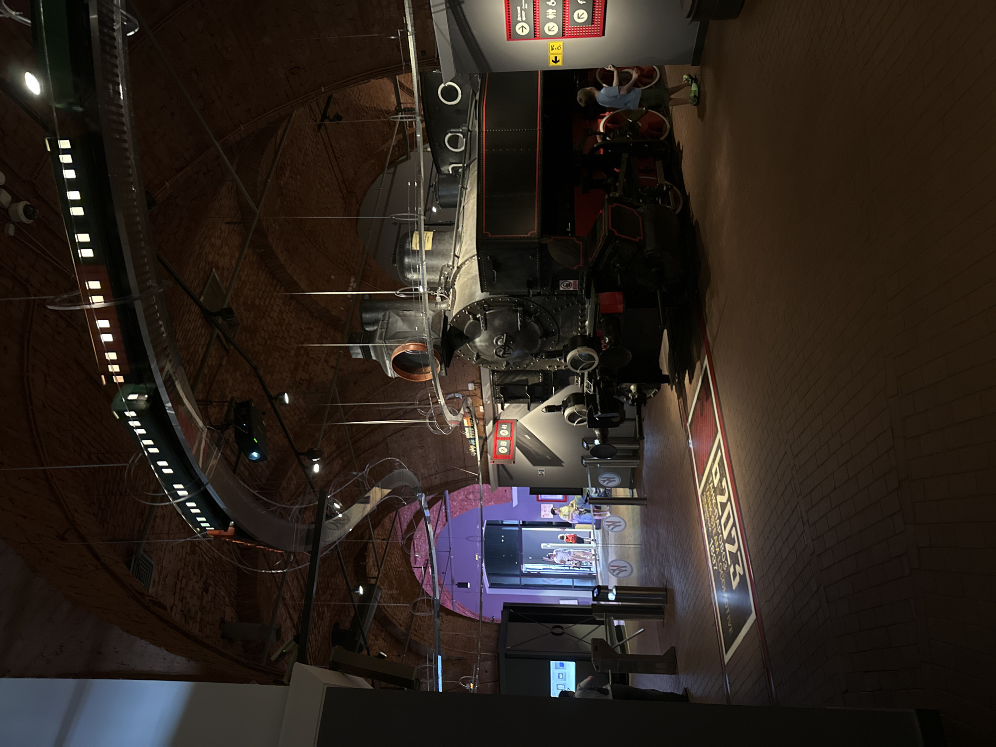 The image depicts the interior of a museum exhibit, likely the Central Naval Museum in Saint Petersburg, Russia, given the presence of historical trains and naval artifacts. In the foreground, a large, black and red steam locomotive is the central exhibit. It is positioned on a brick floor and surrounded by display elements. Above the locomotive, a model train is suspended on a curved track, illuminated and appearing to be in motion.
To the left of the locomotive, there are turnstiles and glass partitions, suggesting an entrance or exit area. A screen displaying information is also visible. Further back, through an arched entryway illuminated with purple light, several visitors can be seen, including a family with children.
To the right of the locomotive, a young boy in a blue t-shirt and shorts is interacting with a display panel. The panel features various signs, including directional arrows and icons related to facilities like a children's center and restrooms. The overall environment suggests a well-lit, spacious exhibition hall with a focus on transportation history. The architecture of the ceiling, with its brickwork and arched structures, indicates a historical building repurposed for the museum. A projected graphic on the floor near the turnstiles displays text and graphics related to "B 2023 TANK STEAM LOCOMOTIVE 1897."