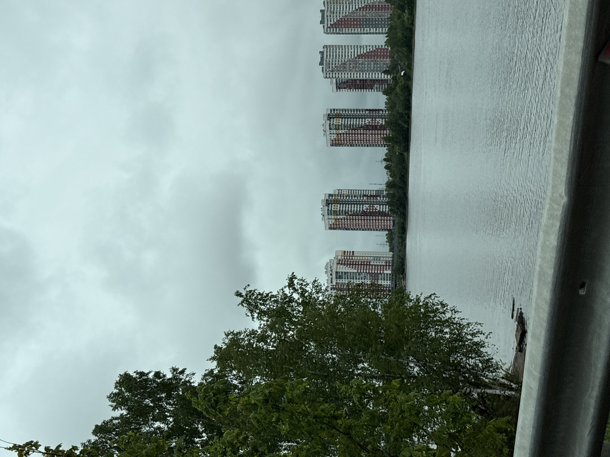 The scene is a view of the river near Krasnaya Zarya, Russia, on an overcast day. In the background are multiple tall, modern apartment buildings lining the opposite riverbank. These buildings are characterized by their white facades and colorful accents in red, orange, and green. The river's surface is calm, showing slight ripples. In the foreground, a section of dark green tree foliage partially obscures the view. A gray metal guardrail runs along the right edge of the image.