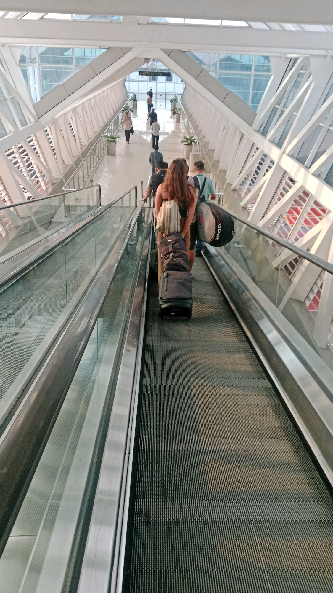 The image shows people on a moving walkway inside an airport terminal in Tangerang, Indonesia. The walkway is designed with a modern, angular architecture, featuring white structural elements and large glass windows. Several individuals are seen moving along the walkway, some carrying luggage. The primary subject in the foreground is a woman with long, dark hair, wearing a striped top and shorts, pulling two large suitcases behind her. To her right, a person with a blue shirt is visible, also carrying luggage. Further up the walkway, other travelers can be seen walking in both directions, with one person in a white shirt and another in a dark shirt. The walkway appears to be indoors, with sunlight filtering through the glass. The overall impression is one of transit and travel within a contemporary airport setting.