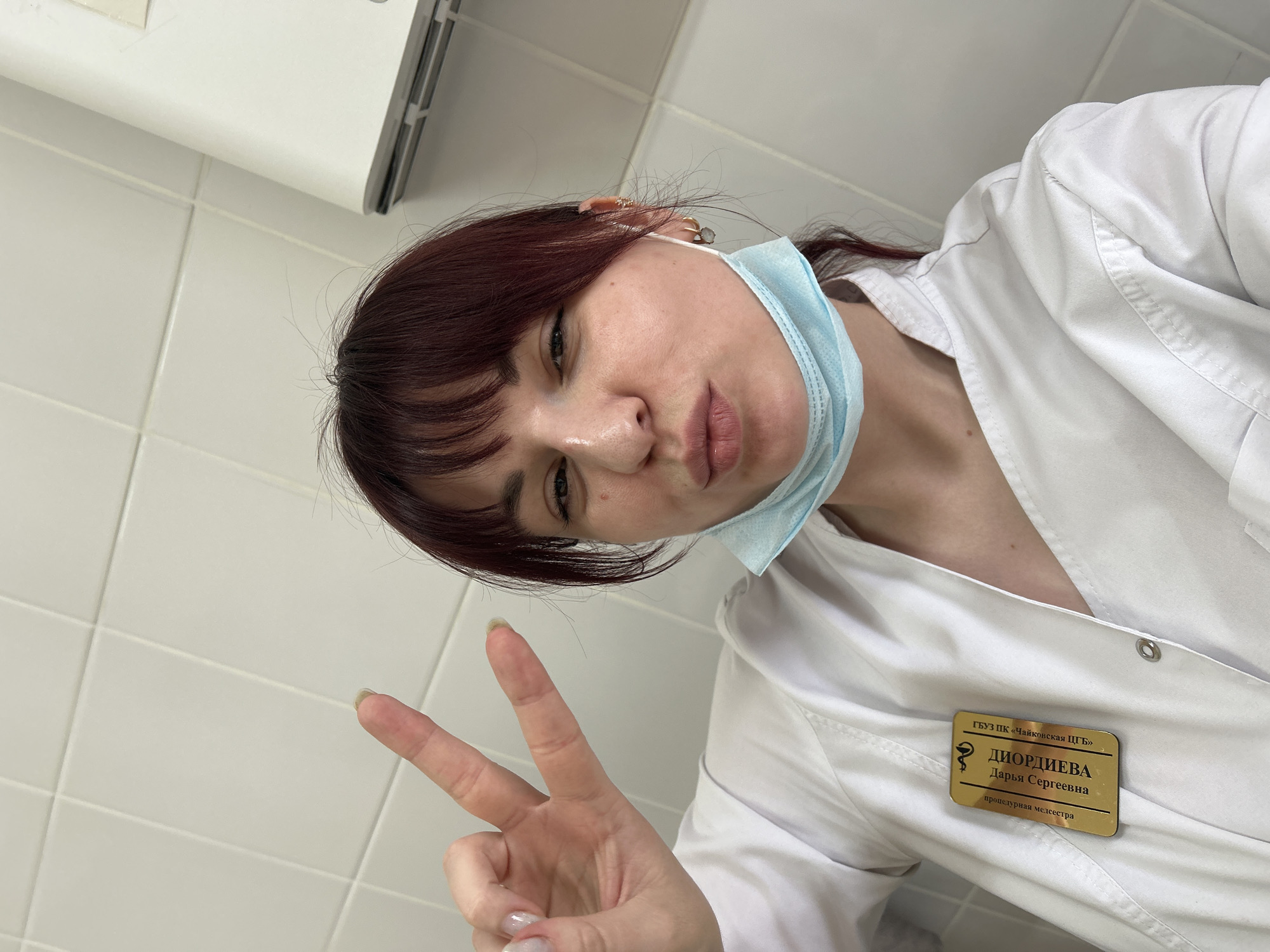 A woman with auburn hair and a surgical mask hanging around her neck poses for a selfie in what appears to be a medical setting. She is wearing a white lab coat with a name tag that reads "ДИОРДИЕВА Дарья Сергеевна процедурная медсестра," identifying her as Dar'ya Sergeyevna Diordiyeva, a procedural nurse. The name tag also includes "ГБУЗ ПК «Чайковская ШГБ»," indicating the location as the Chaykovsky City Hospital in Фоки, Russia. She is holding up a peace sign. The background shows white tiled walls and a white cabinet.