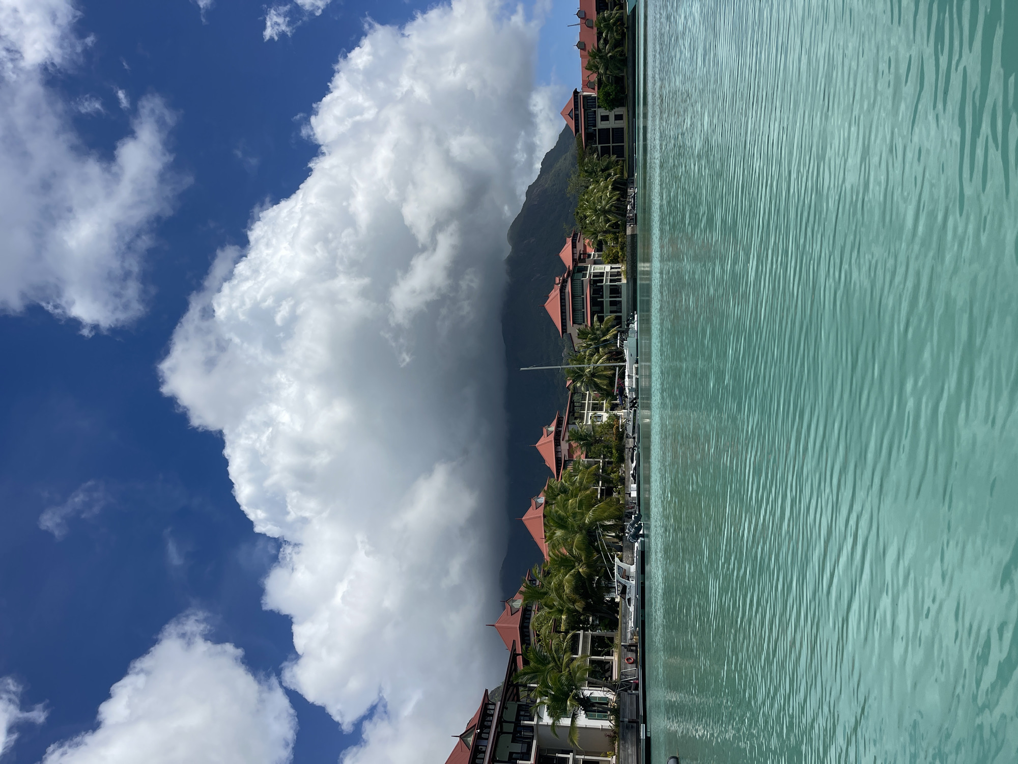 A wide shot captures a serene waterfront scene in Providence, Seychelles, under a bright blue sky dotted with fluffy white clouds. The foreground is dominated by calm, turquoise water with subtle ripples. Beyond the water, a marina is lined with several boats docked along a pier. Behind the marina, a row of multi-story buildings with distinctive red-tiled roofs and white facades stretches across the midground. These buildings are interspersed with lush green palm trees. In the background, a large mountain range is partially obscured by a dramatic, dark cloud formation. The overall atmosphere is tranquil and tropical.