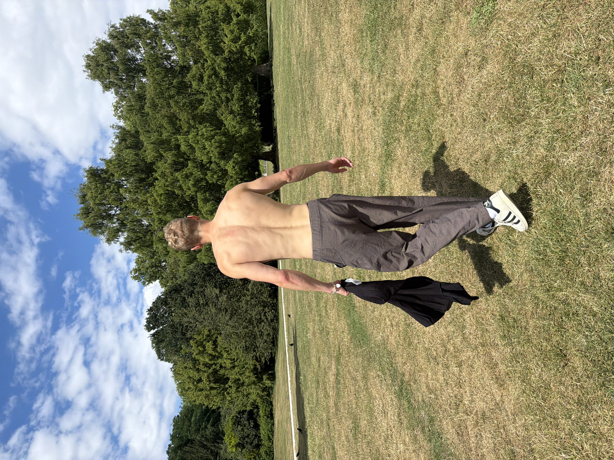 A shirtless man with blonde hair walks away from the camera across a dry, grassy field. He is holding a black t-shirt in his left hand. He is wearing checkered brown pants and white sneakers with black stripes. In the background, there are several large green trees. The sky is blue with scattered white clouds. There are some birds on the grass in the mid-ground. The location is a park, likely in London, United Kingdom.