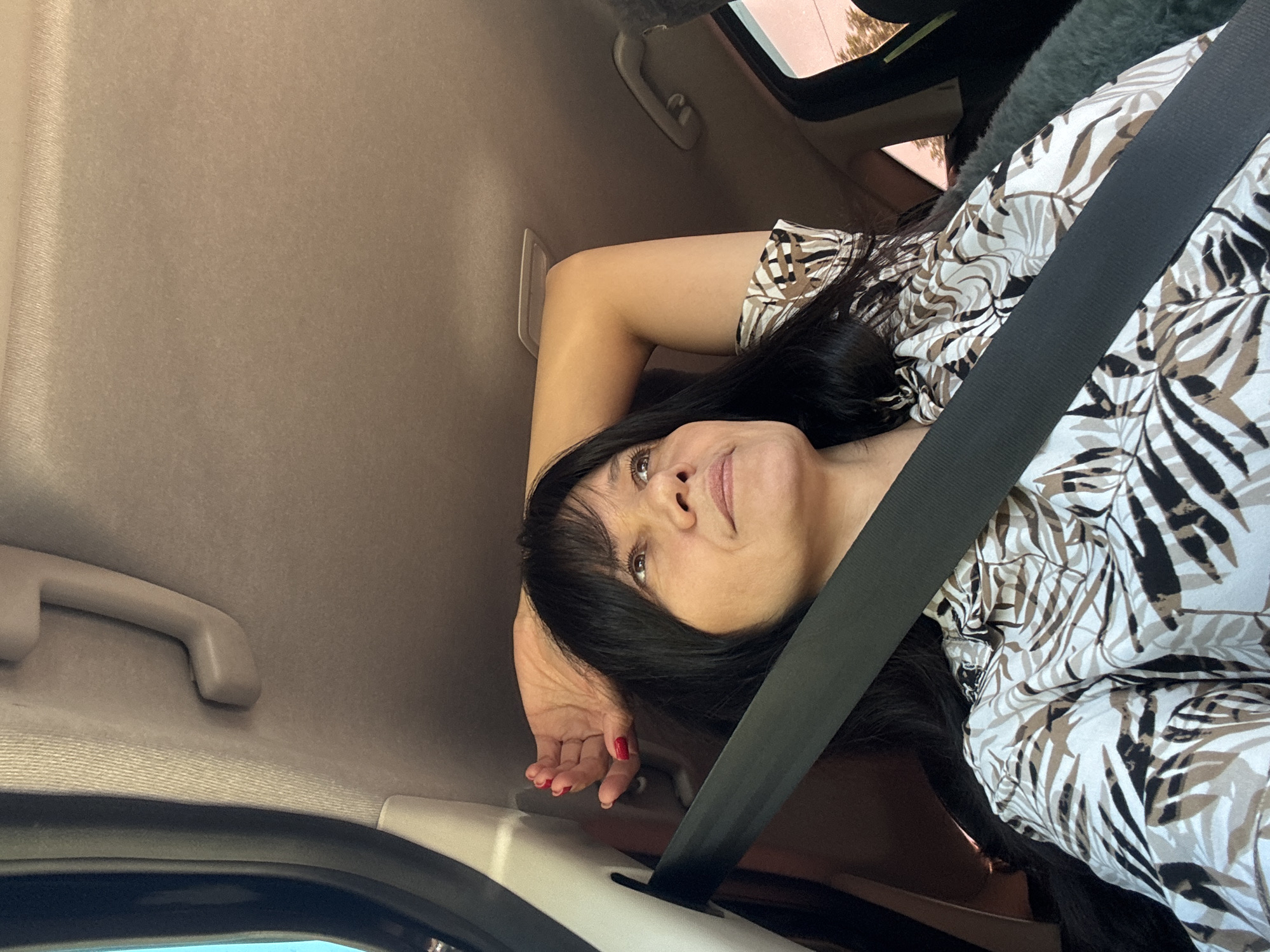 The image shows a woman in the front passenger seat of a car. She is wearing a seatbelt and a patterned shirt or dress with a foliage motif. Her dark hair is styled with bangs. Her left arm is bent with her hand resting on the car interior near the door. Her nails are painted red. The car's interior is beige or tan. Part of the window and a handle are visible. The location is unknown.