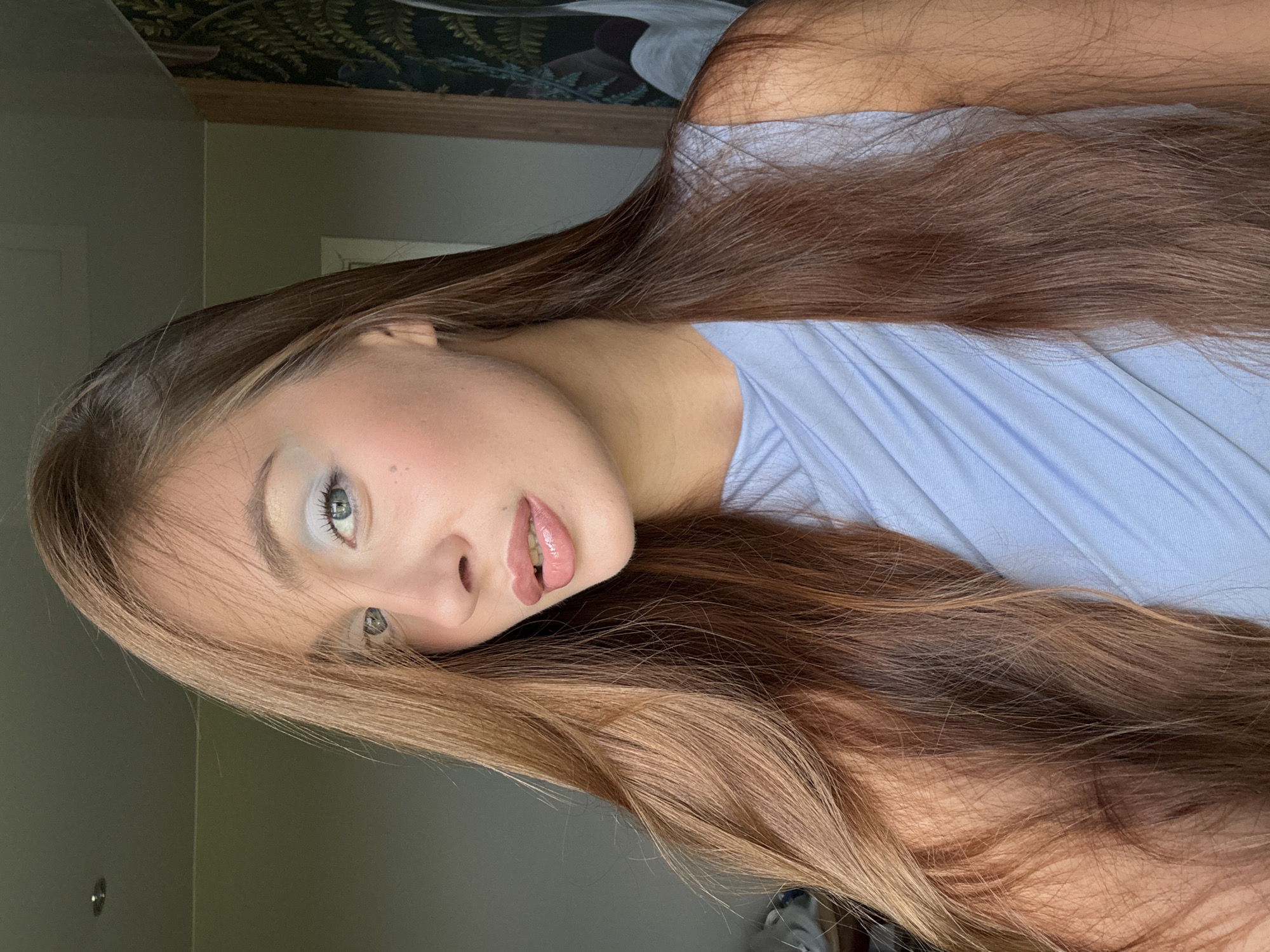 The media file contains a close-up, low-angle portrait of a young person. The subject has long, wavy, brown hair and is wearing a light blue top. Their face is turned towards the camera, with visible makeup on their eyes and lips. The background is out of focus and appears to be an indoor setting, possibly a room with a wall and a framed artwork or tapestry. The lighting suggests it is daytime. There is no visible text in the image. The individual's gaze is directed at the camera, creating an intimate and engaging portrait. The overall mood is serene and contemplative.